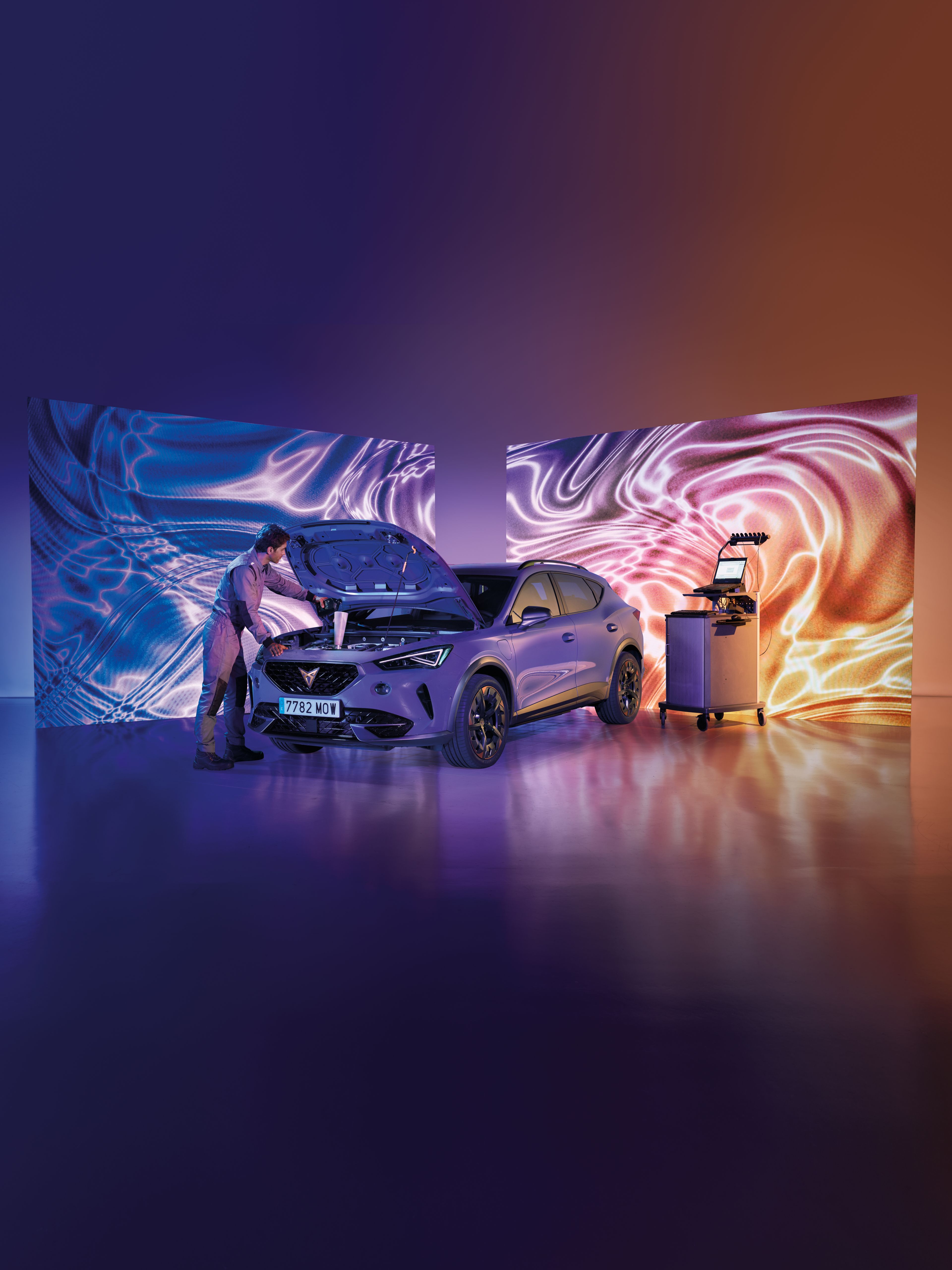 Mechanik kontroluje motor auta v modernom prostredí so svetelnými efektmi na dvoch veľkých obrazovkách. Napravo od auta sa nachádza diagnostický prístroj. Auto je v popredí, obklopené farebnými svetlami.