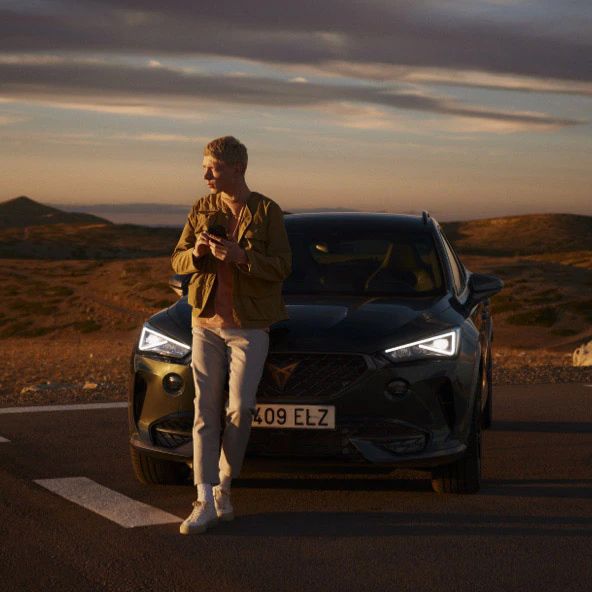 Muž sa opiera o auto na parkovisku v prírodnom prostredí, okolo nej je pustá krajina a na oblohe pomaly zapadá slnko.