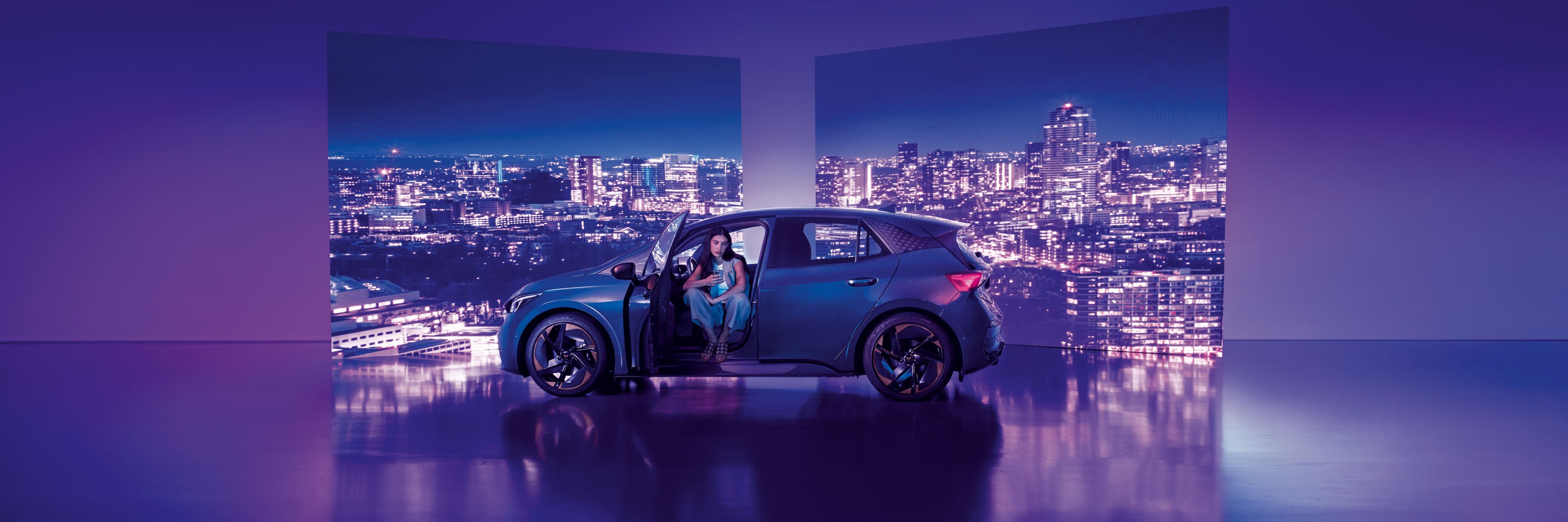 Pozadie tvoria dva veľké billboardy s panoramatickým výhľadom na nočné mesto, pred ktorými stojí moderné auto s otvorenými dverami. Žena sedí v aute na mieste vodiča, interiér je osvetlený jemným svetlom.