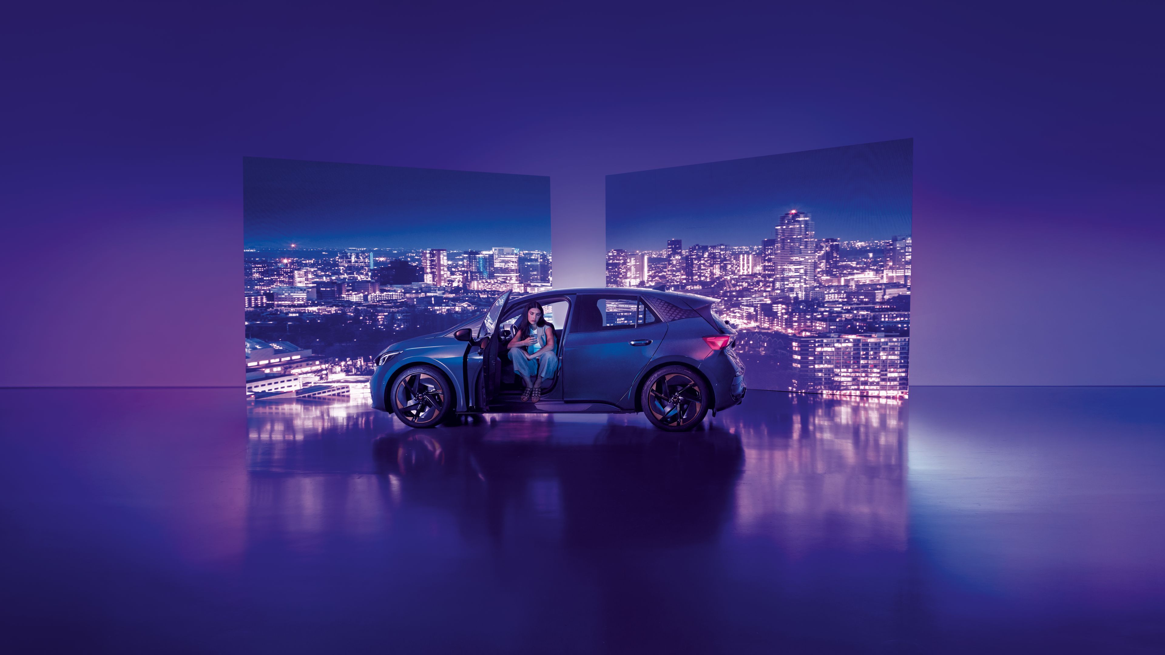 Pozadie tvoria dva veľké billboardy s panoramatickým výhľadom na nočné mesto, pred ktorými stojí moderné auto s otvorenými dverami. Žena sedí v aute na mieste vodiča, interiér je osvetlený jemným svetlom.