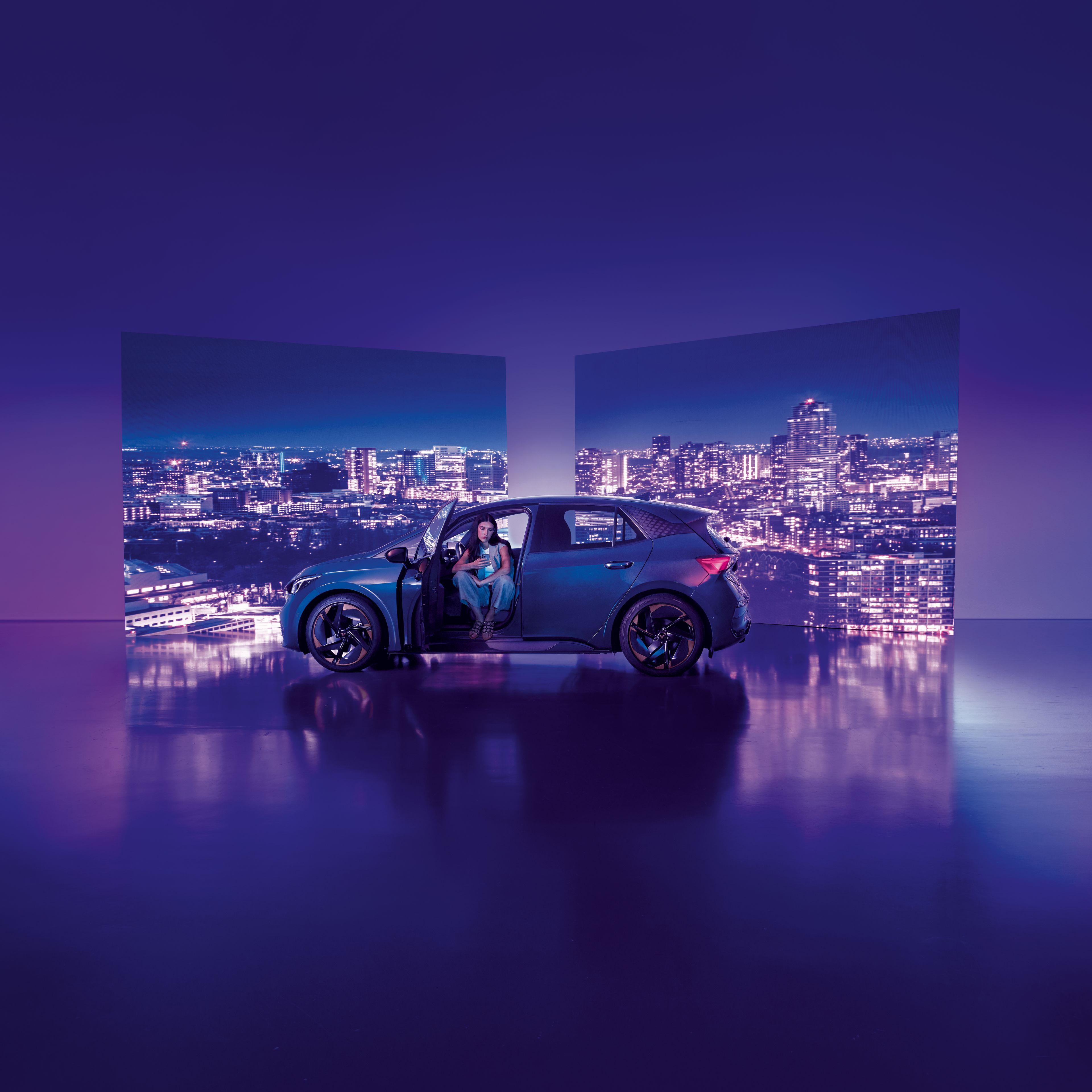 Pozadie tvoria dva veľké billboardy s panoramatickým výhľadom na nočné mesto, pred ktorými stojí moderné auto s otvorenými dverami. Žena sedí v aute na mieste vodiča, interiér je osvetlený jemným svetlom.