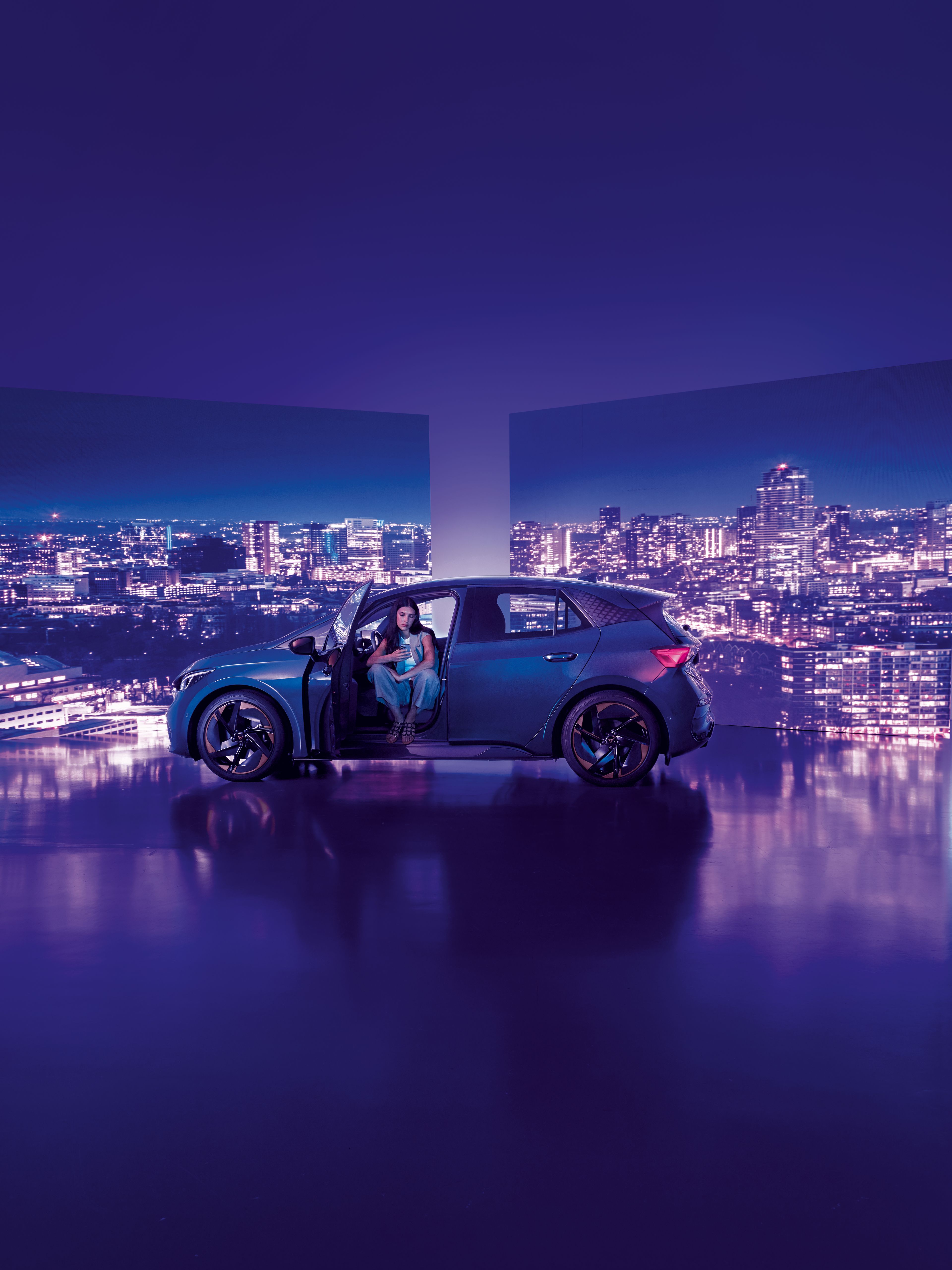 Pozadie tvoria dva veľké billboardy s panoramatickým výhľadom na nočné mesto, pred ktorými stojí moderné auto s otvorenými dverami. Žena sedí v aute na mieste vodiča, interiér je osvetlený jemným svetlom.