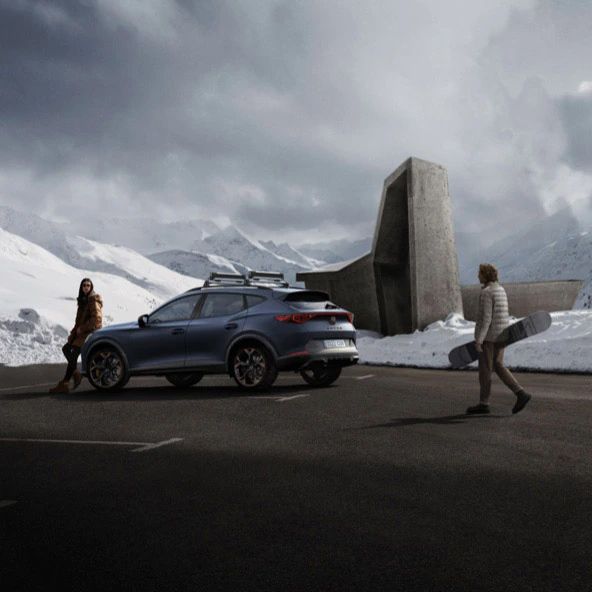 Na obrázku sú dve osoby pri zaparkovanom aute na horskej ceste. Jedna osoba drží snowboard. V pozadí sú zasnežené hory a moderná betónová stavba.