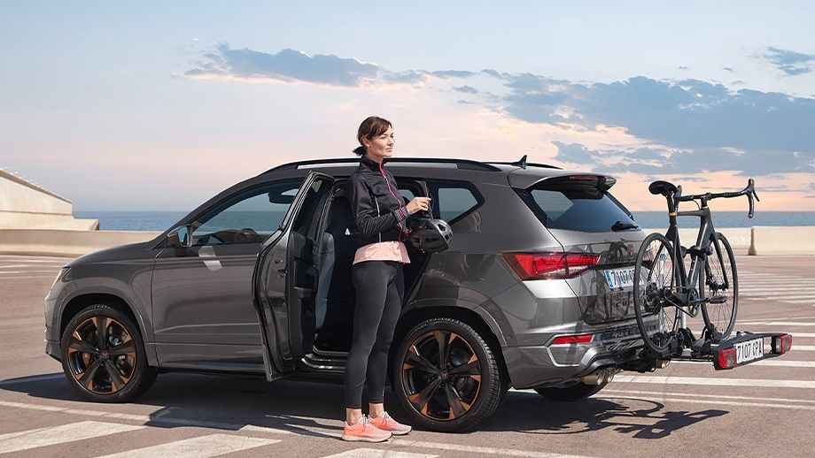 Žena stojí pri strieborno-šedom CUPRA Ateca zaparkovanom na parkovisku. Na zadnej časti auta je upevnený nosič s bicyklom na ťažnom zariadení. Scéna je von, pravdepodobne pri mori alebo oceáne.