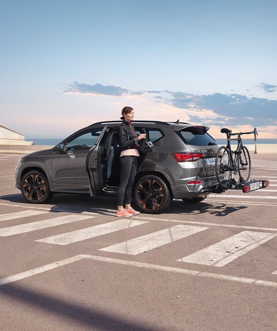 Žena stojí pri strieborno-šedom CUPRA Ateca zaparkovanom na parkovisku. Na zadnej časti auta je upevnený nosič s bicyklom na ťažnom zariadení. Scéna je von, pravdepodobne pri mori alebo oceáne.