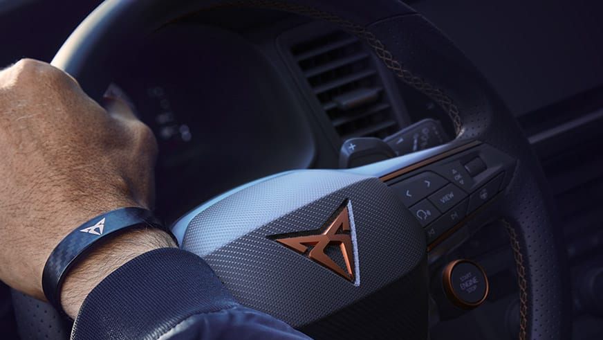 Ruka vodiča na volante auta značky CUPRA. Na zápästí má tmavo modrý náramok s logom CUPRA.