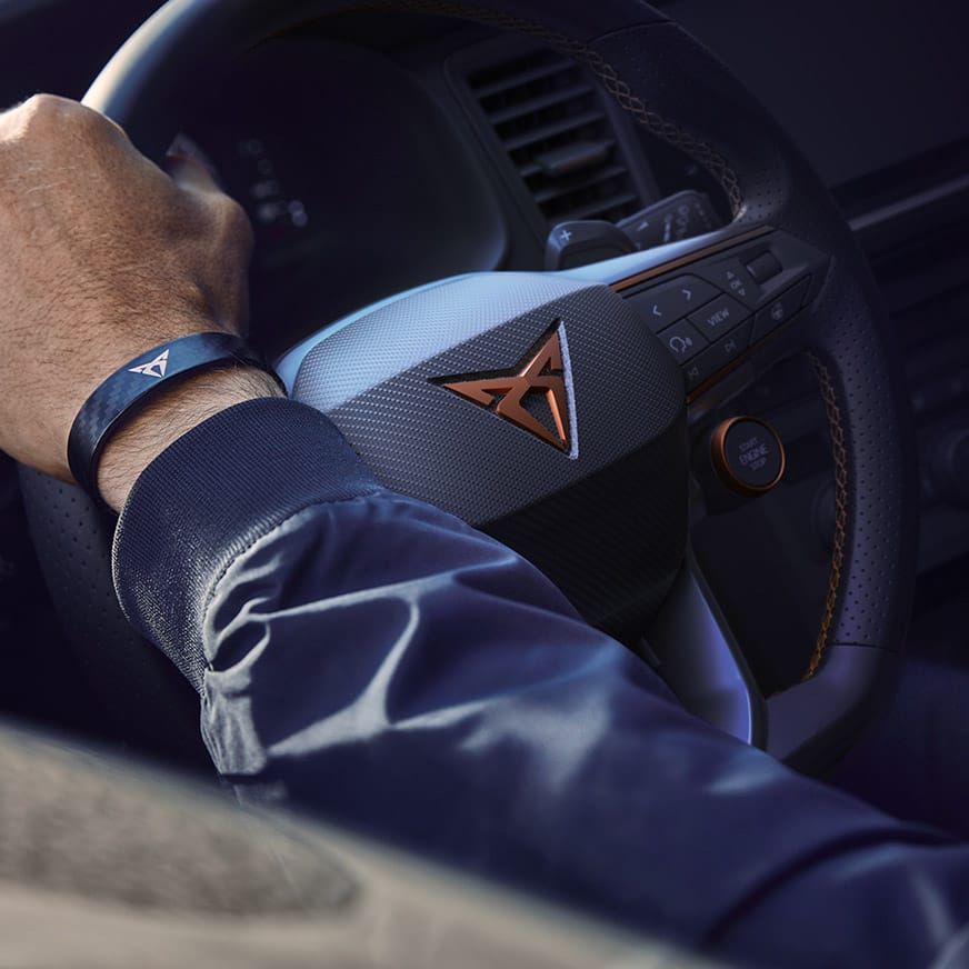 Ruka vodiča na volante auta značky CUPRA. Na zápästí má tmavo modrý náramok s logom CUPRA.