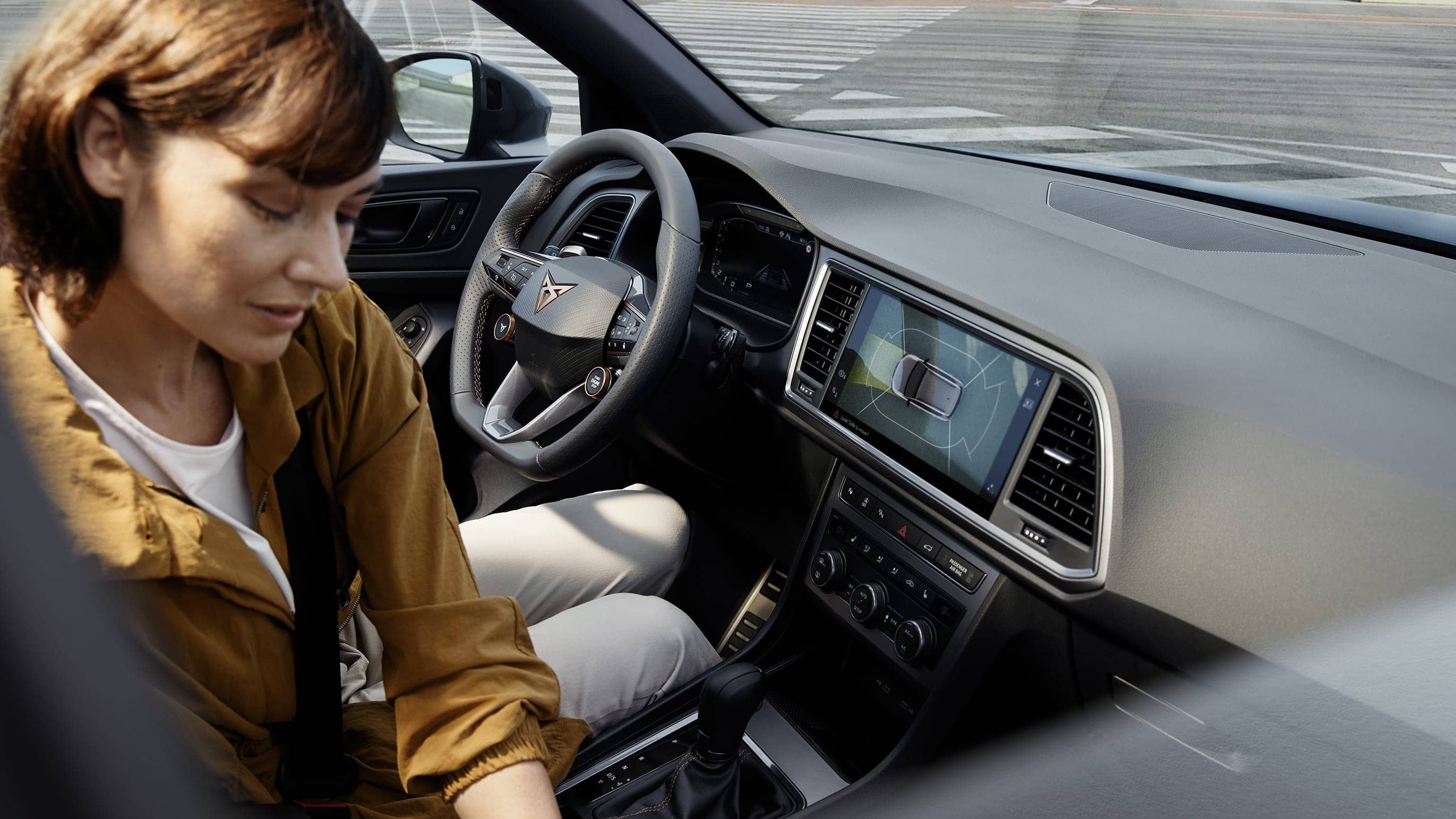 Žena sedí v aute. Vidieť tiež palubnú dosku s moderným dotykovým displejom na ovládanie auta. Interiér vozidla je elegantný a má futuristický dizajn.