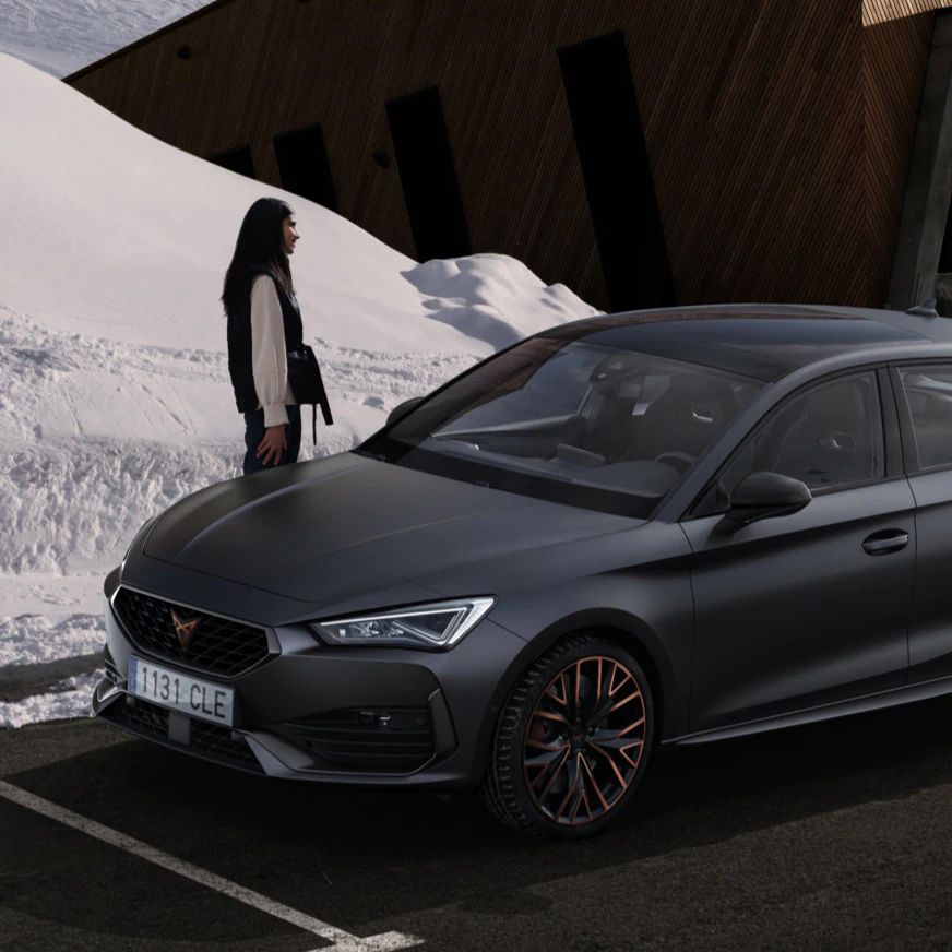 Na obrázku je zaparkované tmavo sivé auto, ktoré stojí pred drevenou budovou pri zasneženom svahu. Vedľa auta stojí osoba. 