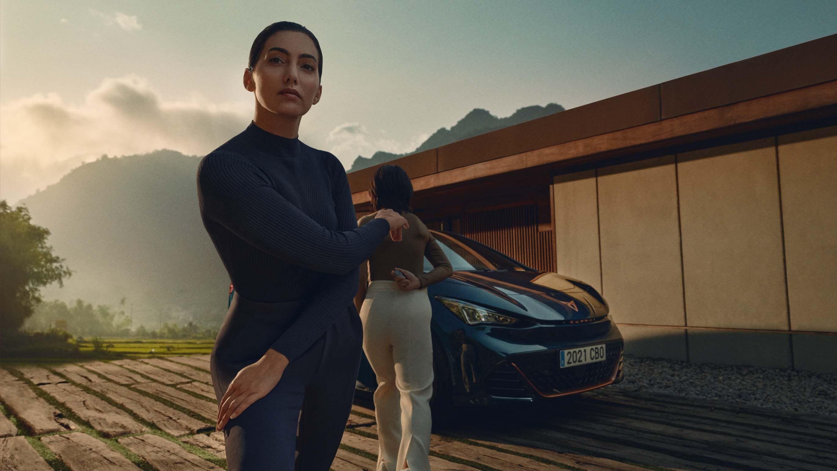 Dve ženy stojace pred luxusným autom s horskou krajinou v pozadí. Automobil má moderný dizajn.