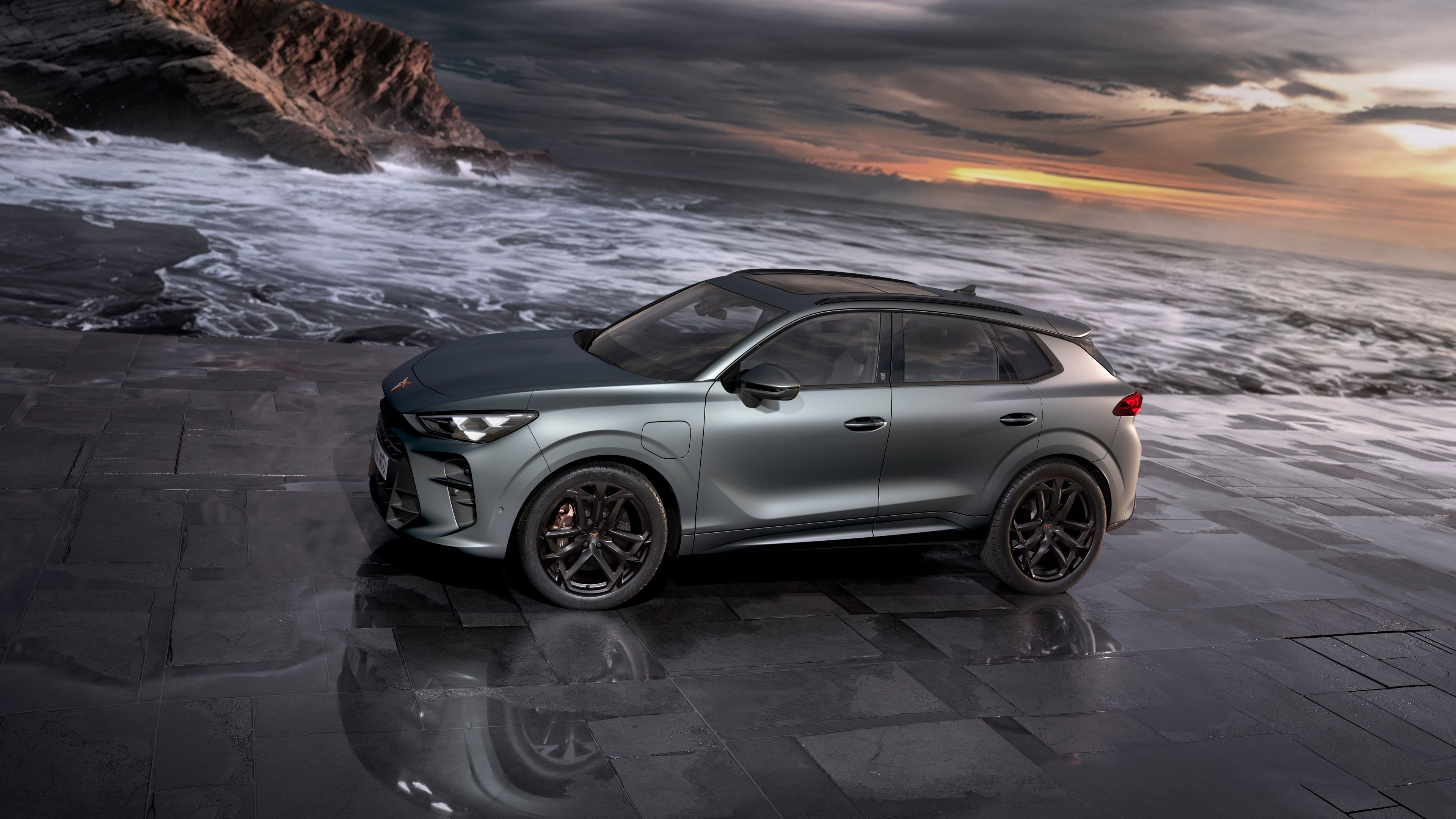 Luxusné športové SUV zaparkované na pobreží s dramatickou oblohou a vlnami rozbíjajúcimi sa o skaly v pozadí. Auto je striebornej farby.