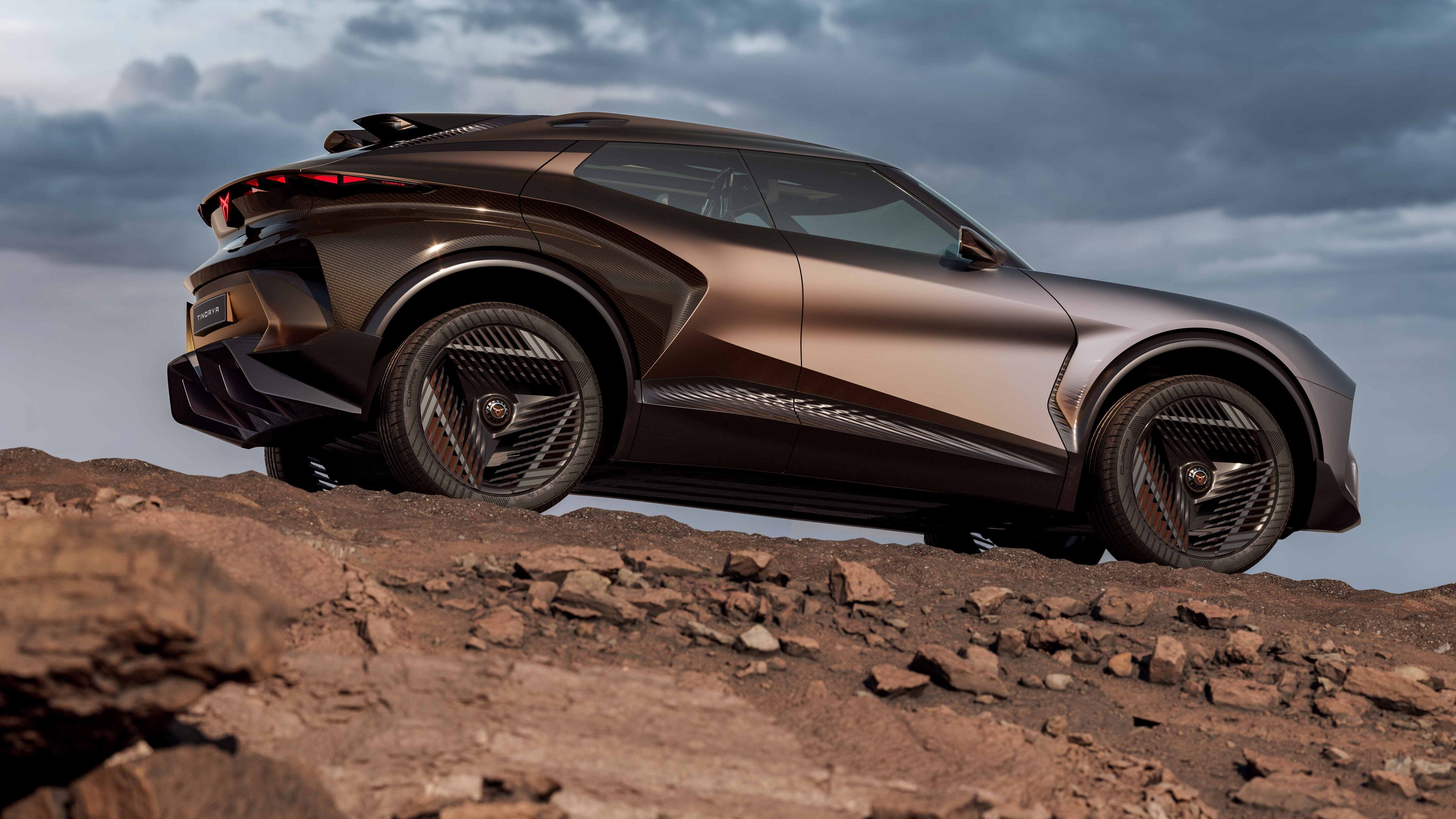 Futuristické športové SUV zaparkované na skalnatom povrchu. Auto má tmavohnedý nádych a moderný, aerodynamický dizajn. Na oblohe sa zhromažďujú temné mraky, čo dodáva dramatickú atmosféru.