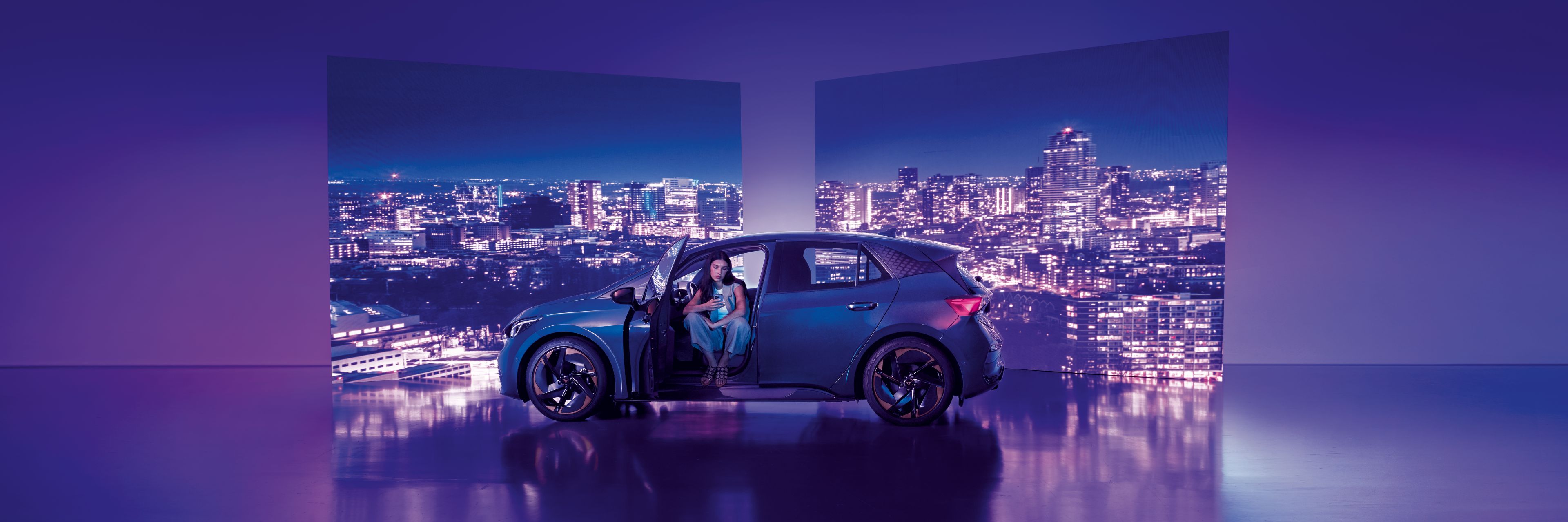 Pozadie tvoria dva veľké billboardy s panoramatickým výhľadom na nočné mesto, pred ktorými stojí moderné auto s otvorenými dverami. Žena sedí v aute na mieste vodiča, interiér je osvetlený jemným svetlom.