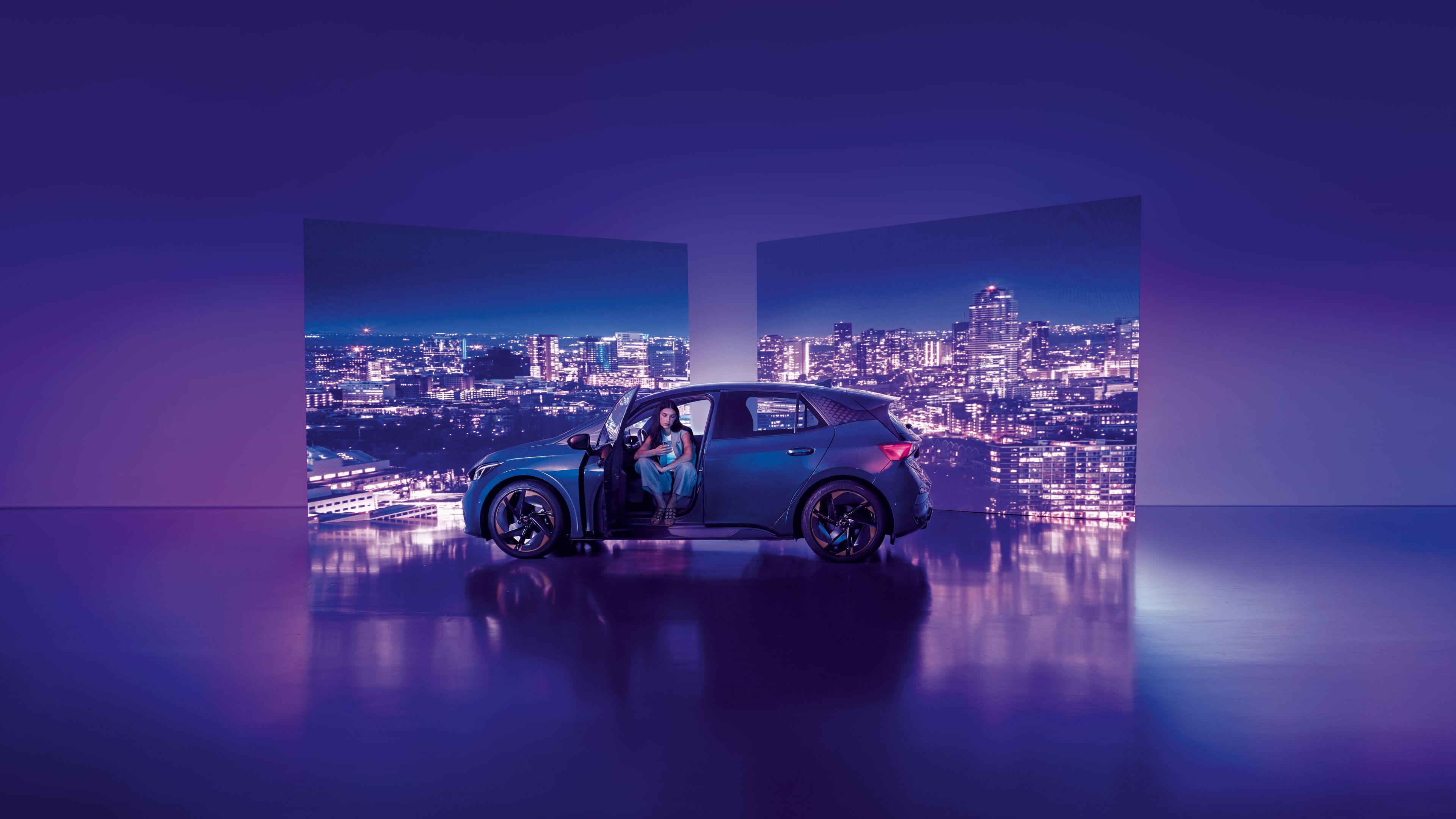Pozadie tvoria dva veľké billboardy s panoramatickým výhľadom na nočné mesto, pred ktorými stojí moderné auto s otvorenými dverami. Žena sedí v aute na mieste vodiča, interiér je osvetlený jemným svetlom.