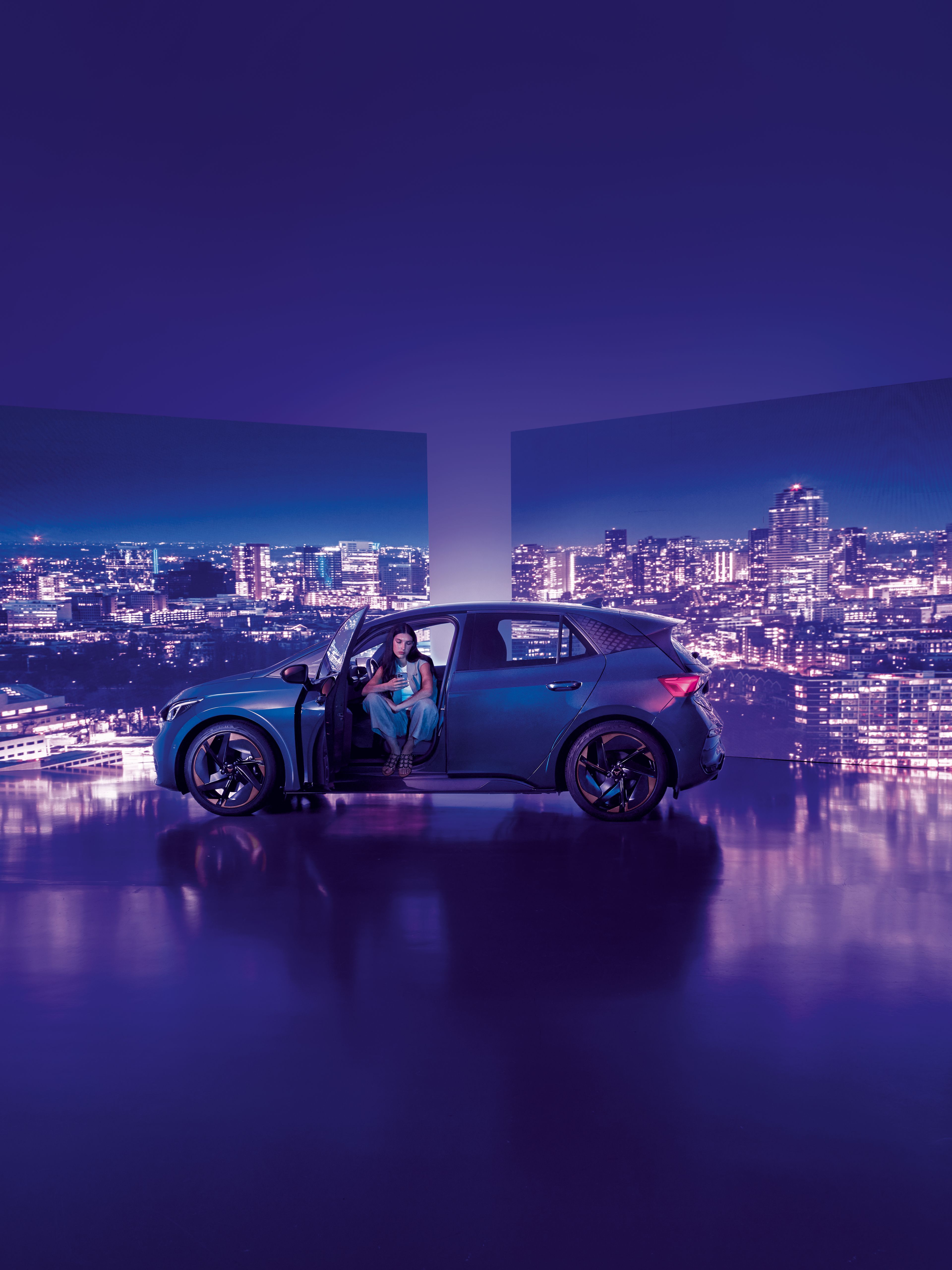 Pozadie tvoria dva veľké billboardy s panoramatickým výhľadom na nočné mesto, pred ktorými stojí moderné auto s otvorenými dverami. Žena sedí v aute na mieste vodiča, interiér je osvetlený jemným svetlom.