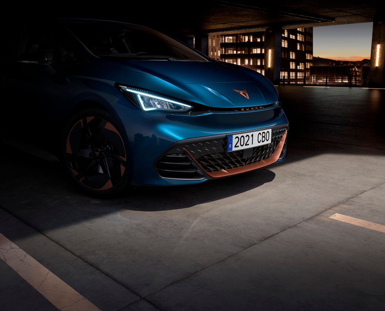 Modré auto zaparkované v garáži s osvetleným mestom v pozadí. Na ŠPZ je uvedený rok 2021. Predné svetlá auta sú zapnuté.