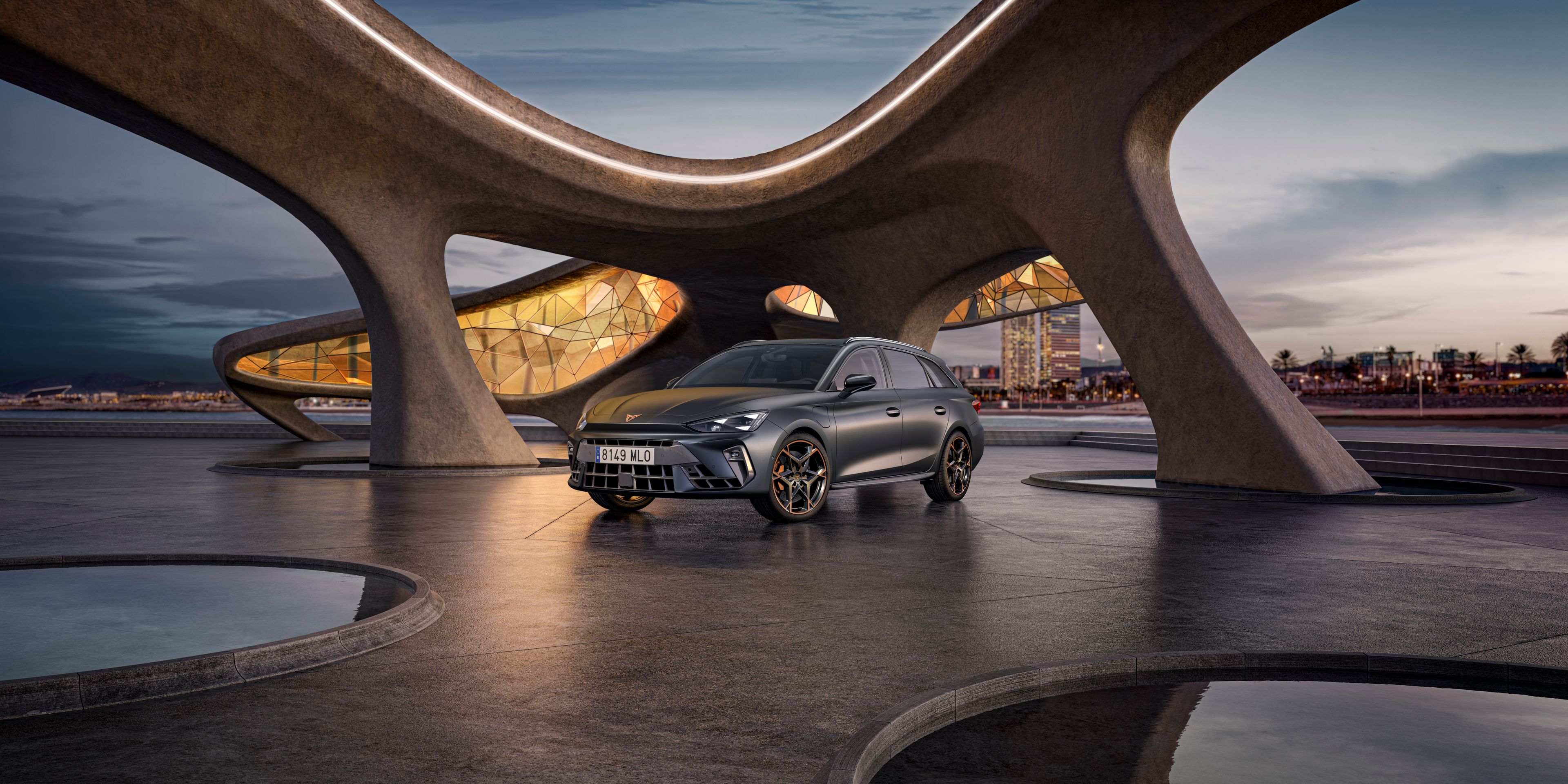 CUPRA Leon Sportstourer na voľnej ploche pri futuristickej budove pri mori. V pozadí je výhľad na nočné mesto.