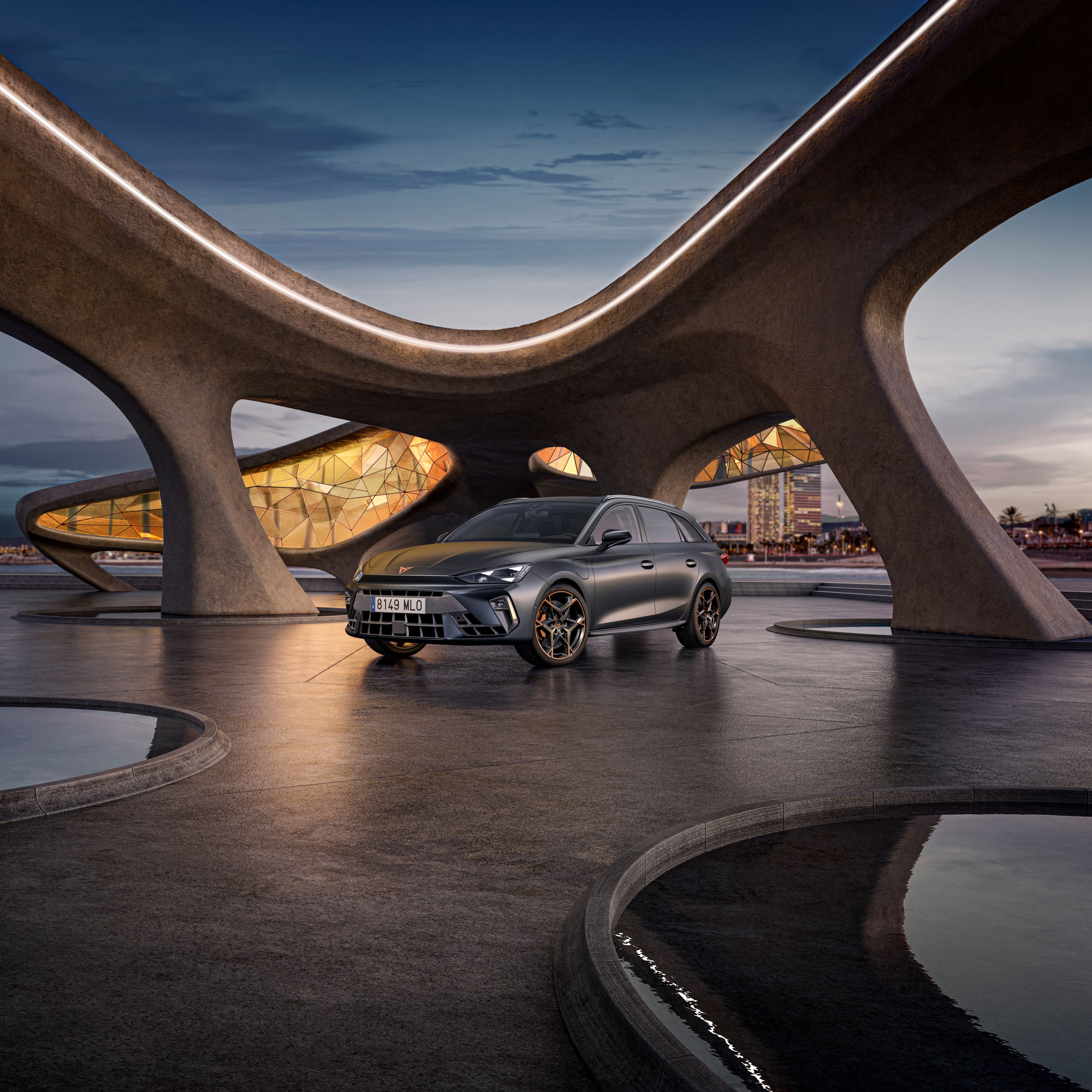 CUPRA Leon Sportstourer na voľnej ploche pri futuristickej budove pri mori. V pozadí je výhľad na nočné mesto.