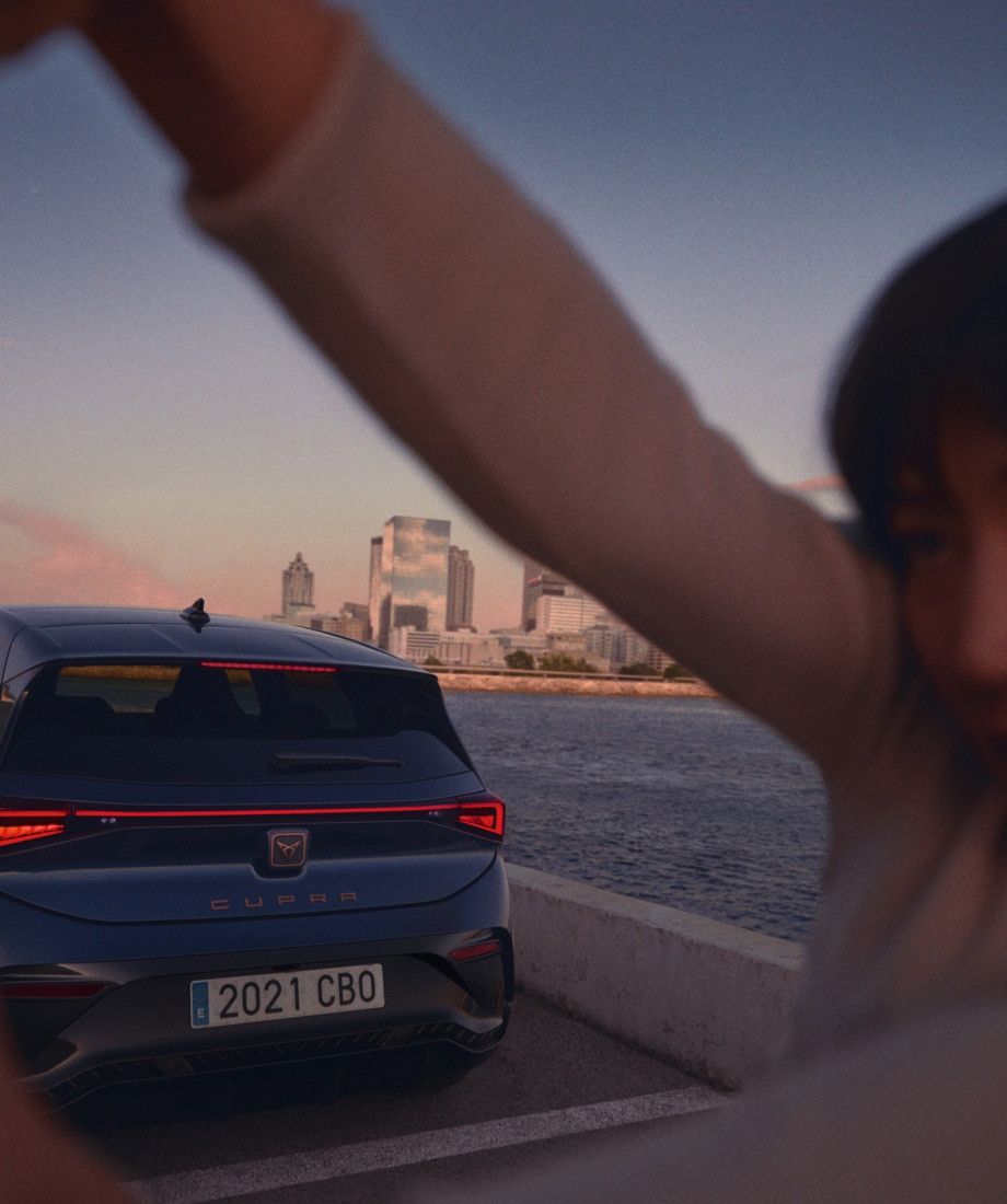 Modré CUPRA Born zaparkované na móle blízko mora, vzadu je panoráma mesta s vysokými budovami. V popredí je rozmazaný obrázok osoby.
