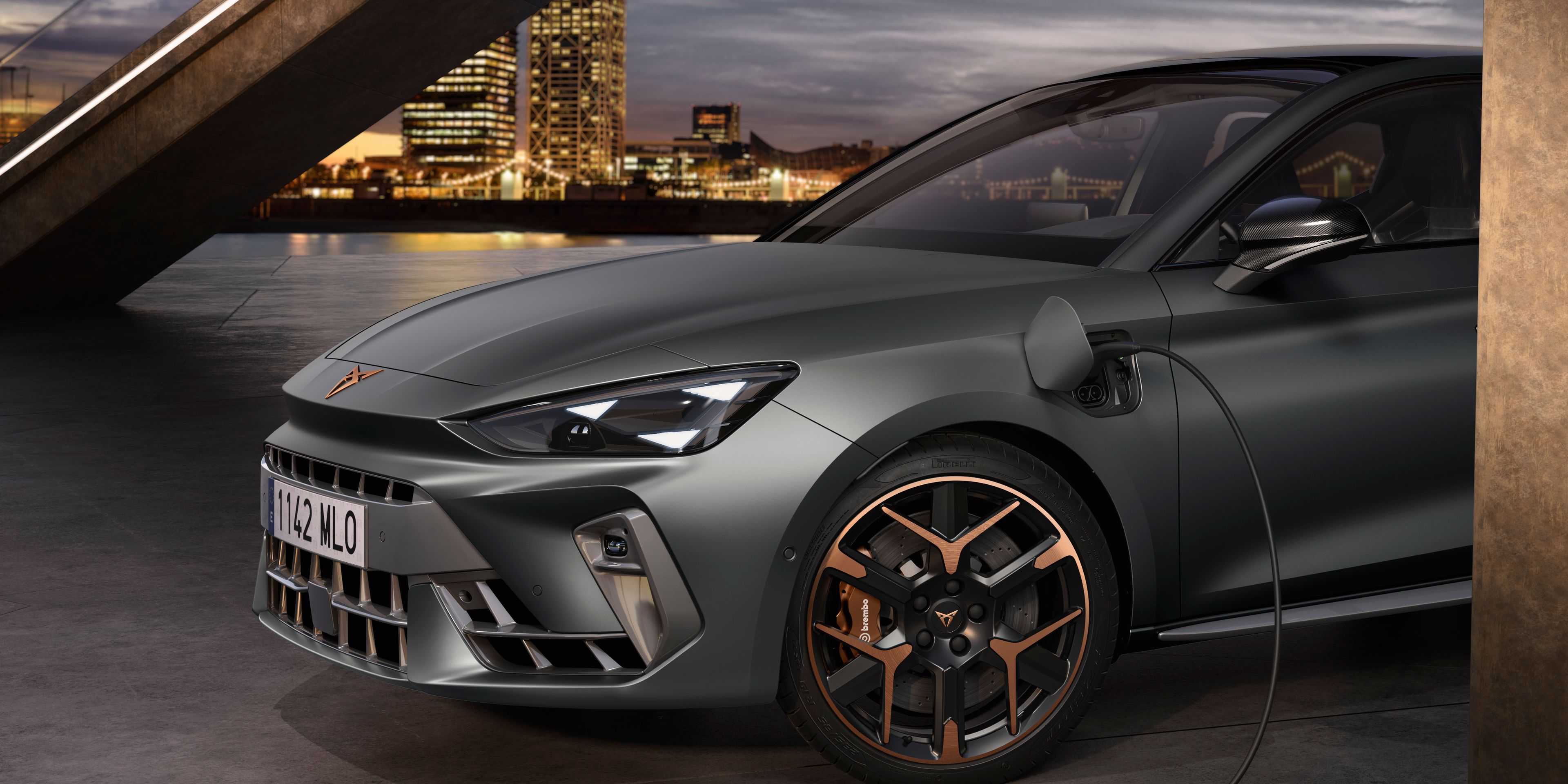 Elektrický automobil Cupra Leon je pripojený k nabíjaciemu káblu v modernom mestskom prostredí. V pozadí je vidieť betónovú lávku pre peších a mrakodrapy. Auto je šedej farby s bronzovými detailmi na kolesách.