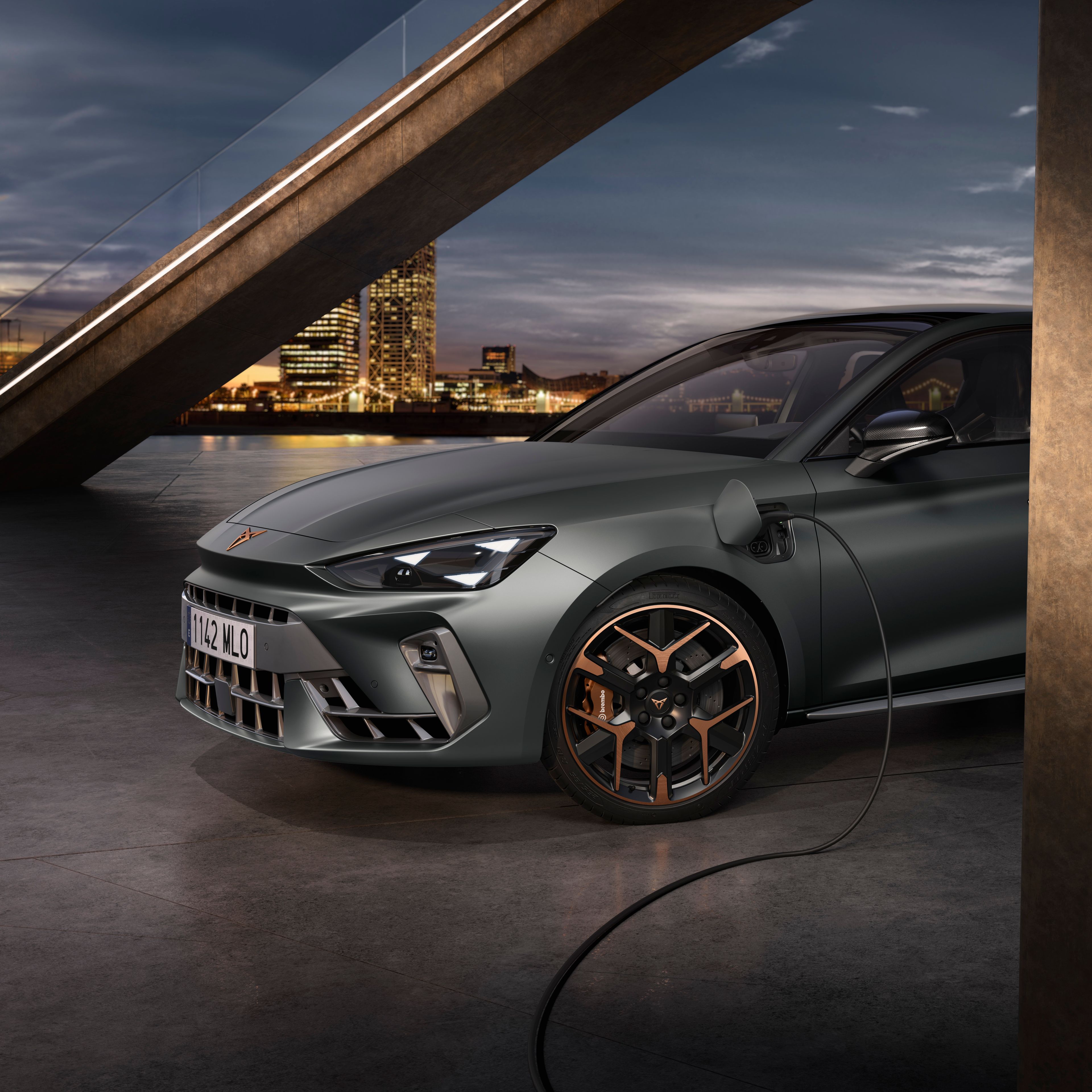 Elektrický automobil Cupra Leon je pripojený k nabíjaciemu káblu v modernom mestskom prostredí. V pozadí je vidieť betónovú lávku pre peších a mrakodrapy. Auto je šedej farby s bronzovými detailmi na kolesách.