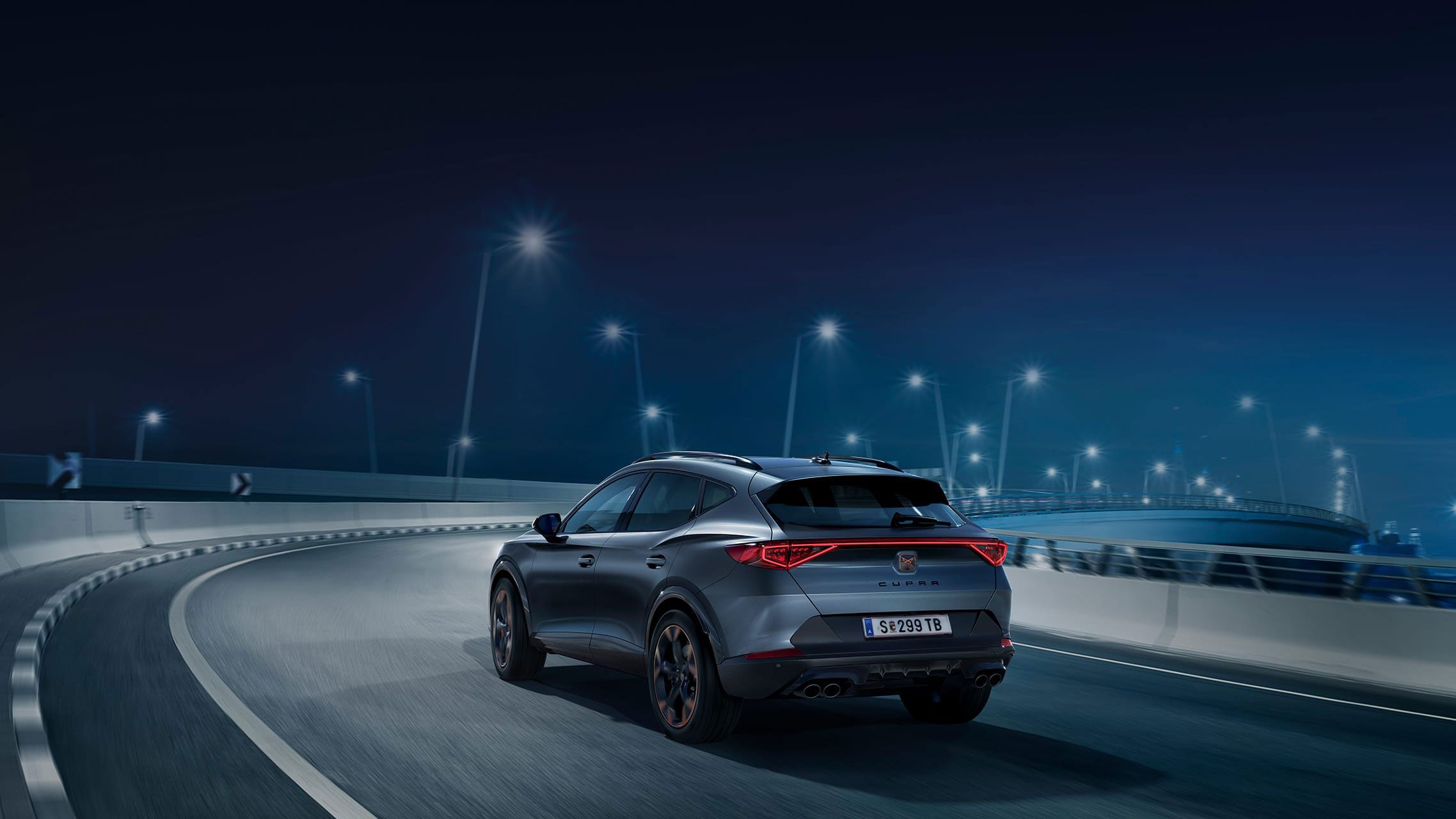 Šedé športové SUV CUPRA jazdí po osvetlenej ceste v noci. Zadná časť vozidla je jasne viditeľná. Okolo auta sú rozmazané svetlá pouličných lámp.