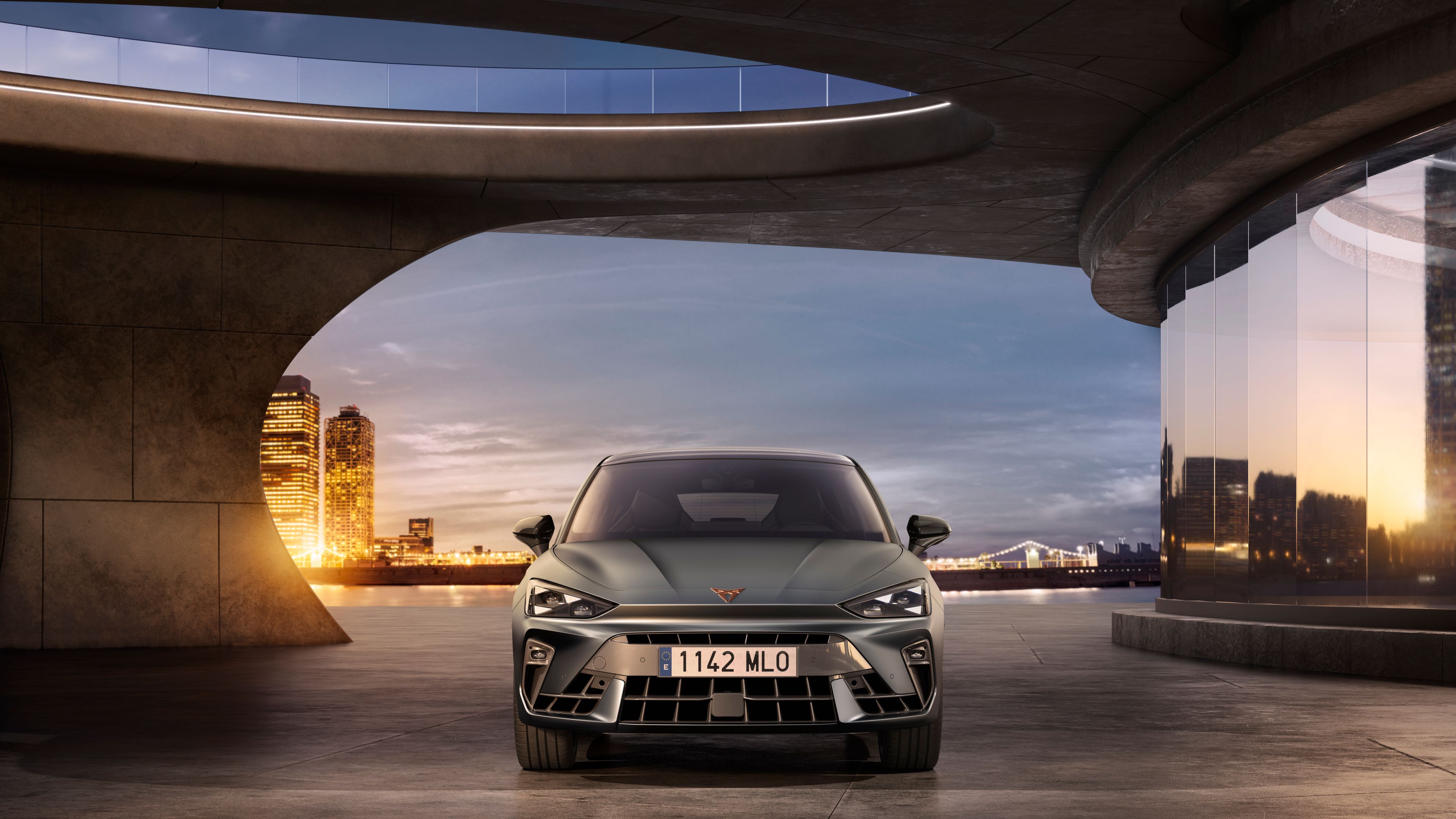 CUPRA Leon zaparkovaný na voľnom priestranstve pri futuristickej budove. V pozadí je výhľad na panorámu nočného mesta.