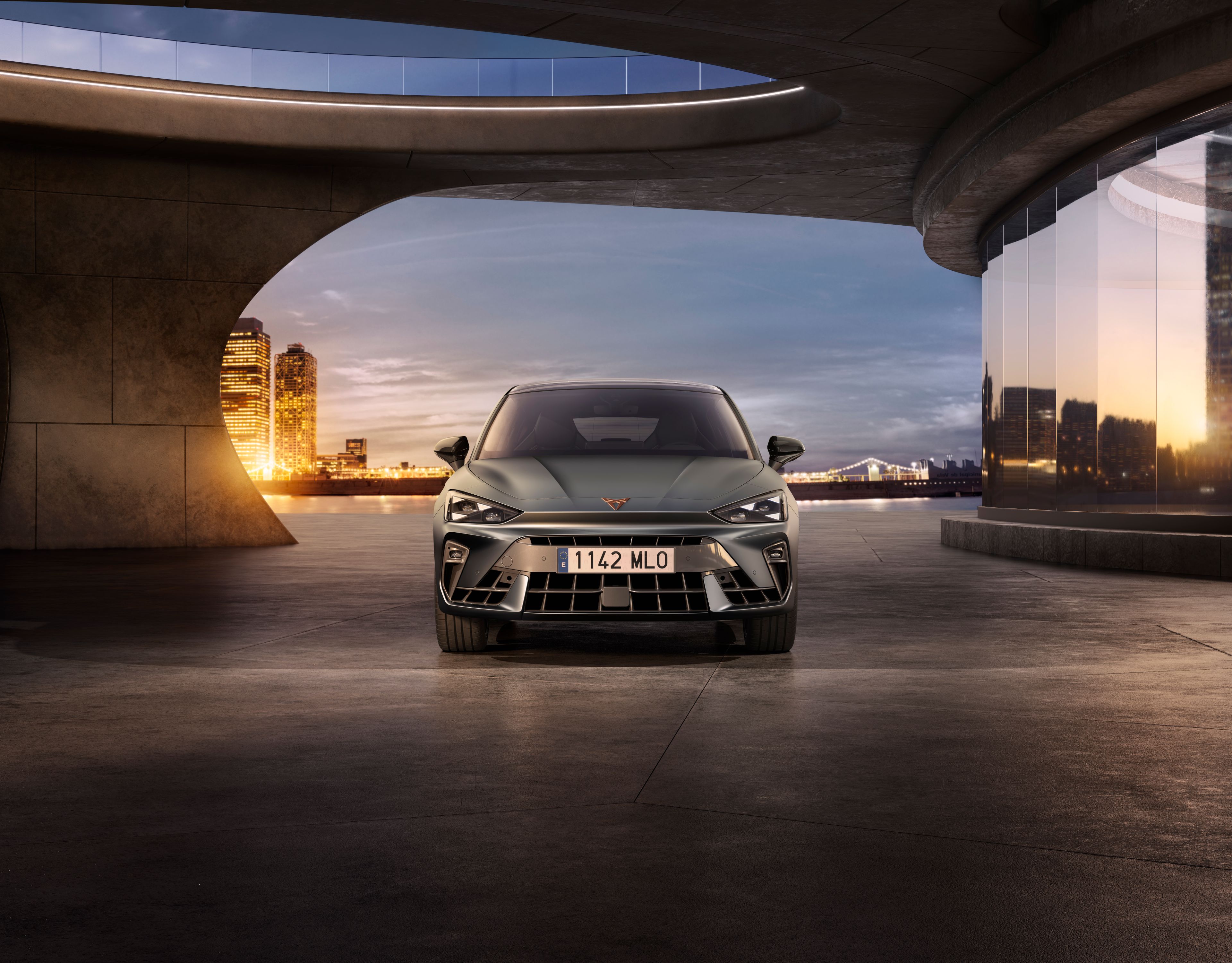 CUPRA Leon zaparkovaný na voľnom priestranstve pri futuristickej budove. V pozadí je výhľad na panorámu nočného mesta.