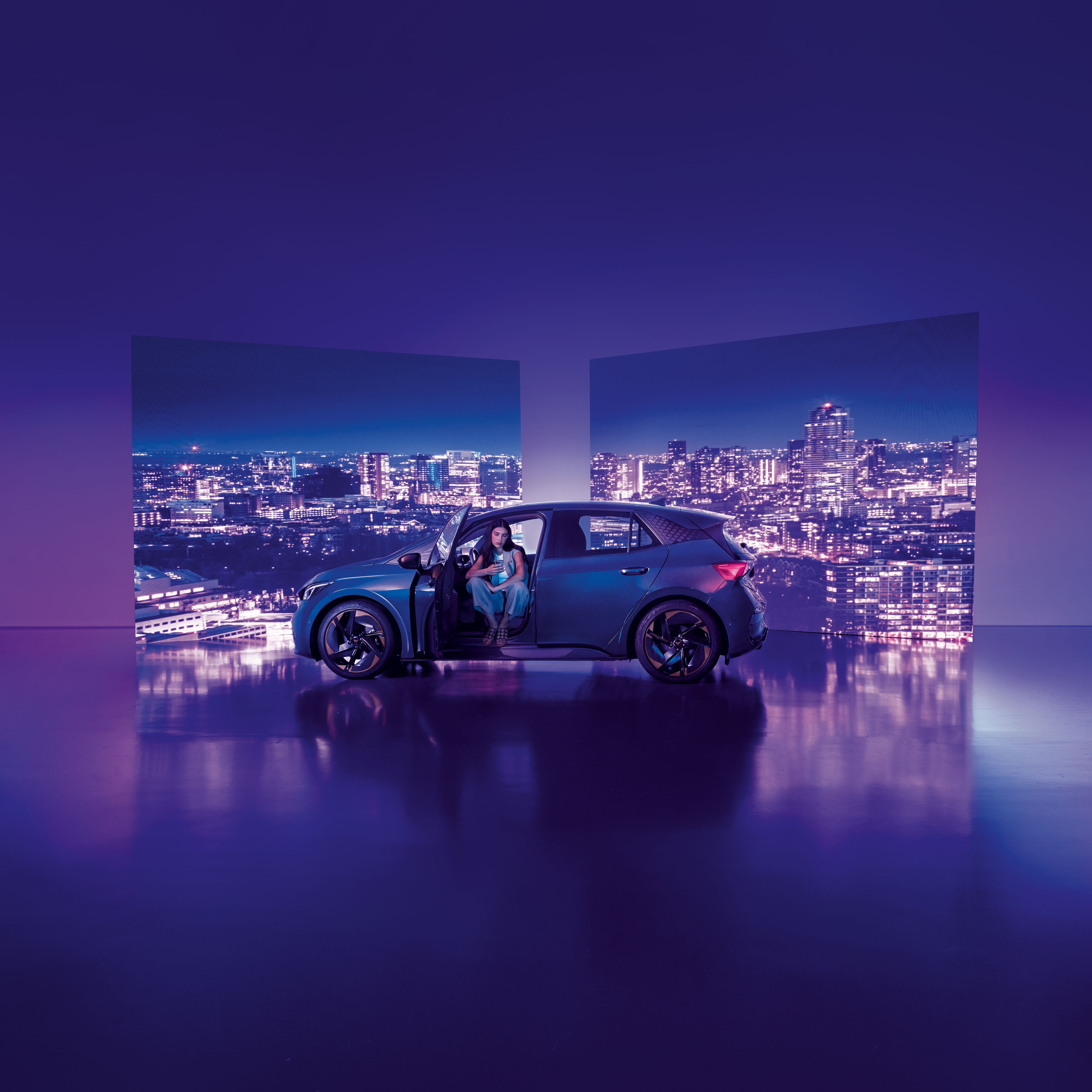 Pozadie tvoria dva veľké billboardy s panoramatickým výhľadom na nočné mesto, pred ktorými stojí moderné auto s otvorenými dverami. Žena sedí v aute na mieste vodiča, interiér je osvetlený jemným svetlom.