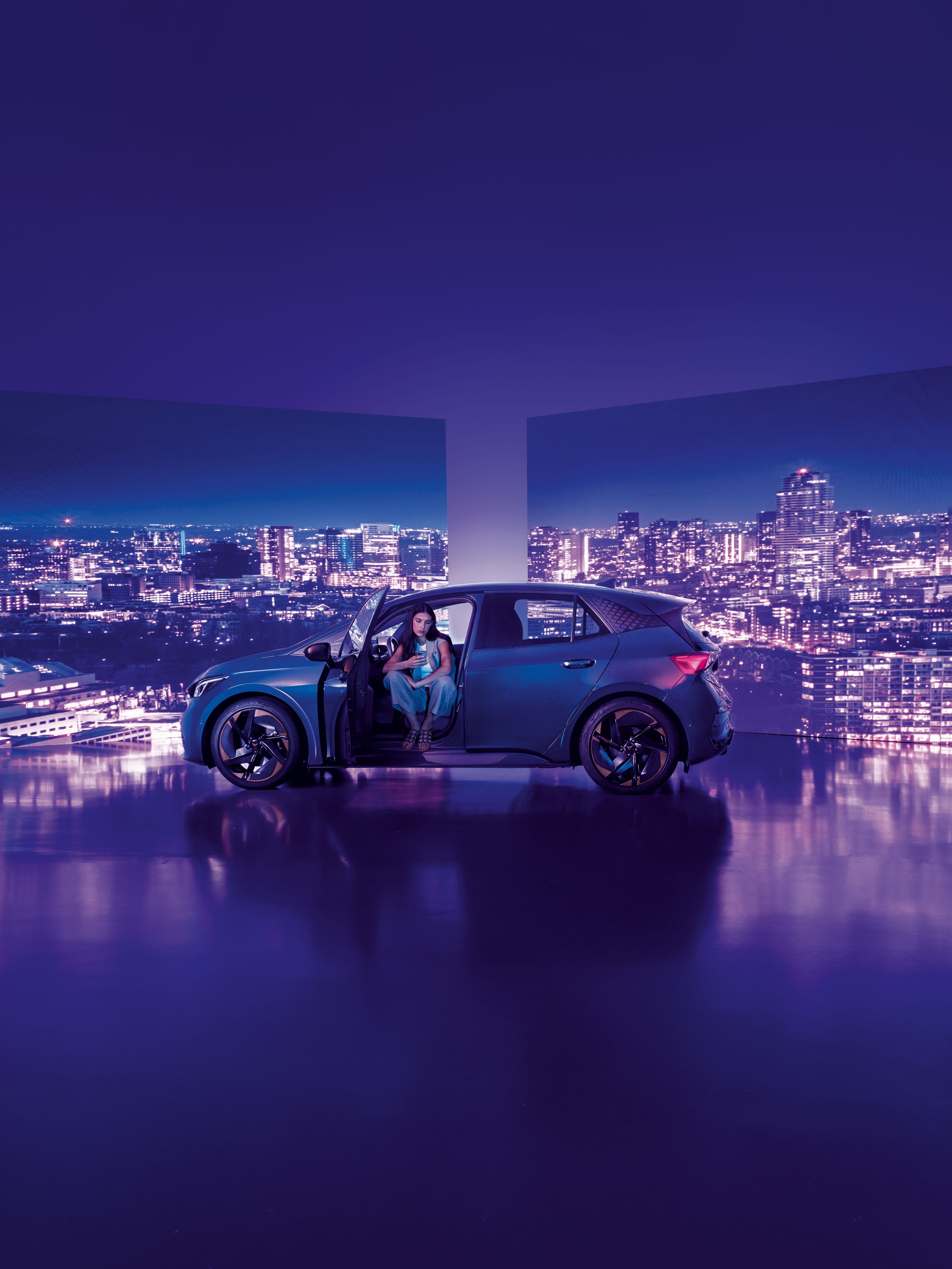 Pozadie tvoria dva veľké billboardy s panoramatickým výhľadom na nočné mesto, pred ktorými stojí moderné auto s otvorenými dverami. Žena sedí v aute na mieste vodiča, interiér je osvetlený jemným svetlom.