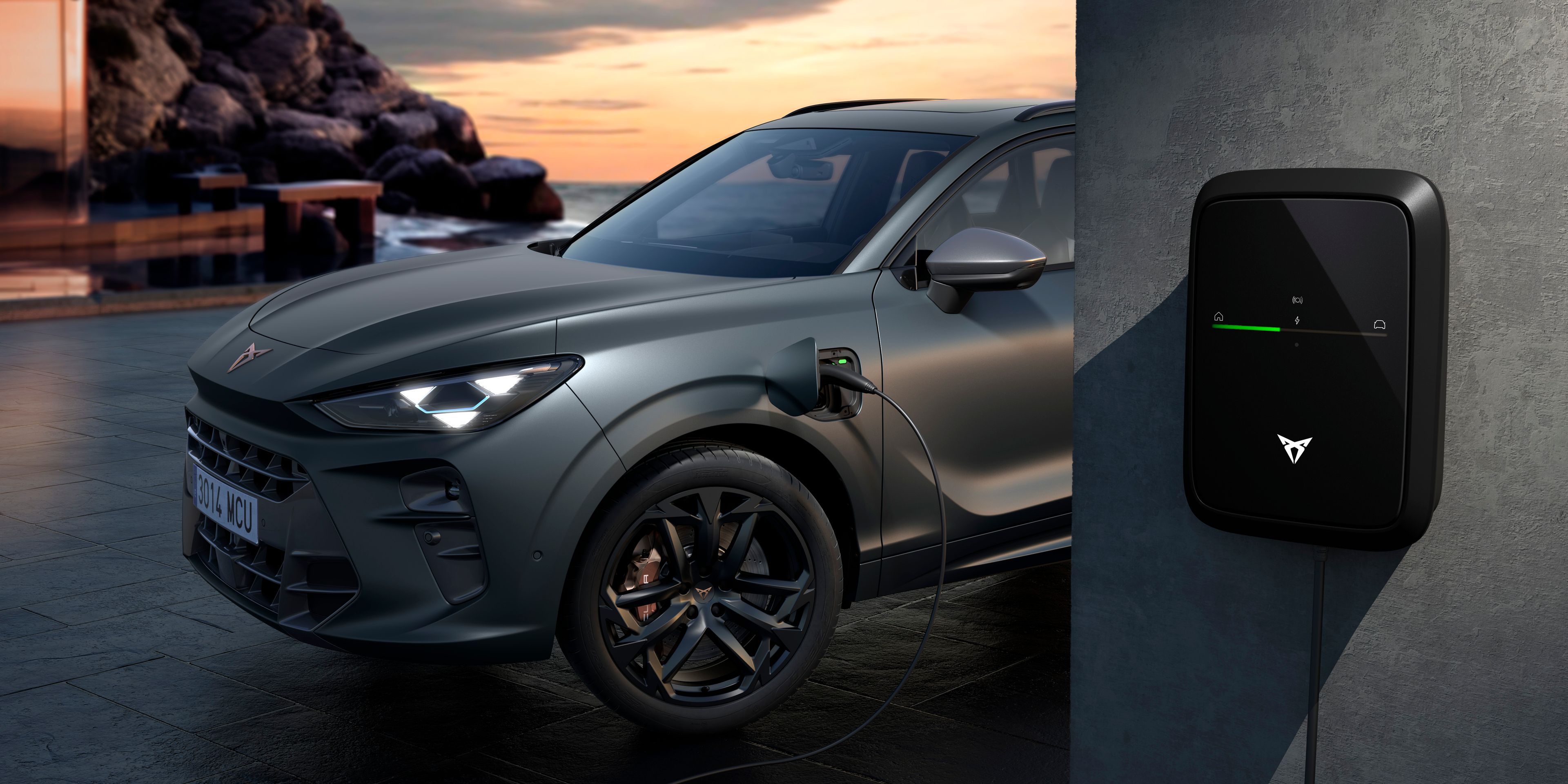 Na obrázku je moderné elektrické SUV, ktoré je pripojené k nabíjacej stanici pri západe slnka. Nabíjacia stanica je upevnená na stene vedľa auta. V pozadí je vidieť elegantnú stavbu a skalnaté pobrežie.