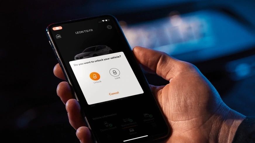 Ruka drží telefón, na ktorom je zobrazená aplikácia s možnosťami odomknutia auta. V pozadí telefónu je rozostrené auto. Upozornenie na displeji ponúka voľbu medzi tlačidlami na odomknutie a uzamknutie vozidla.