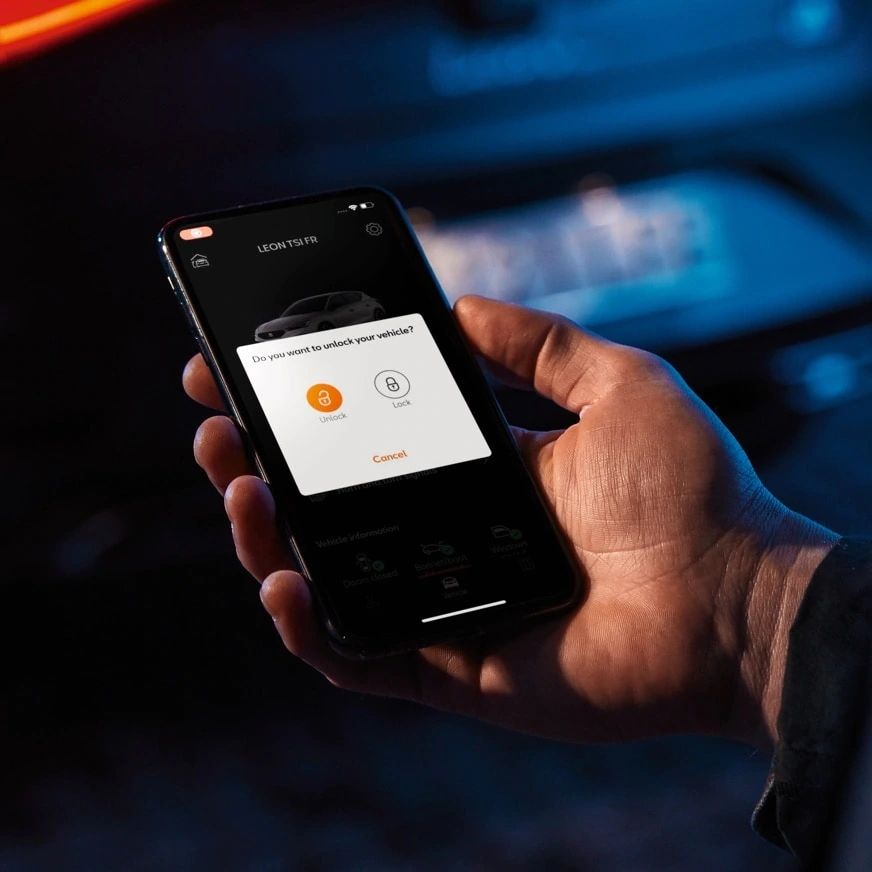 Ruka drží telefón, na ktorom je zobrazená aplikácia s možnosťami odomknutia auta. V pozadí telefónu je rozostrené auto. Upozornenie na displeji ponúka voľbu medzi tlačidlami na odomknutie a uzamknutie vozidla.