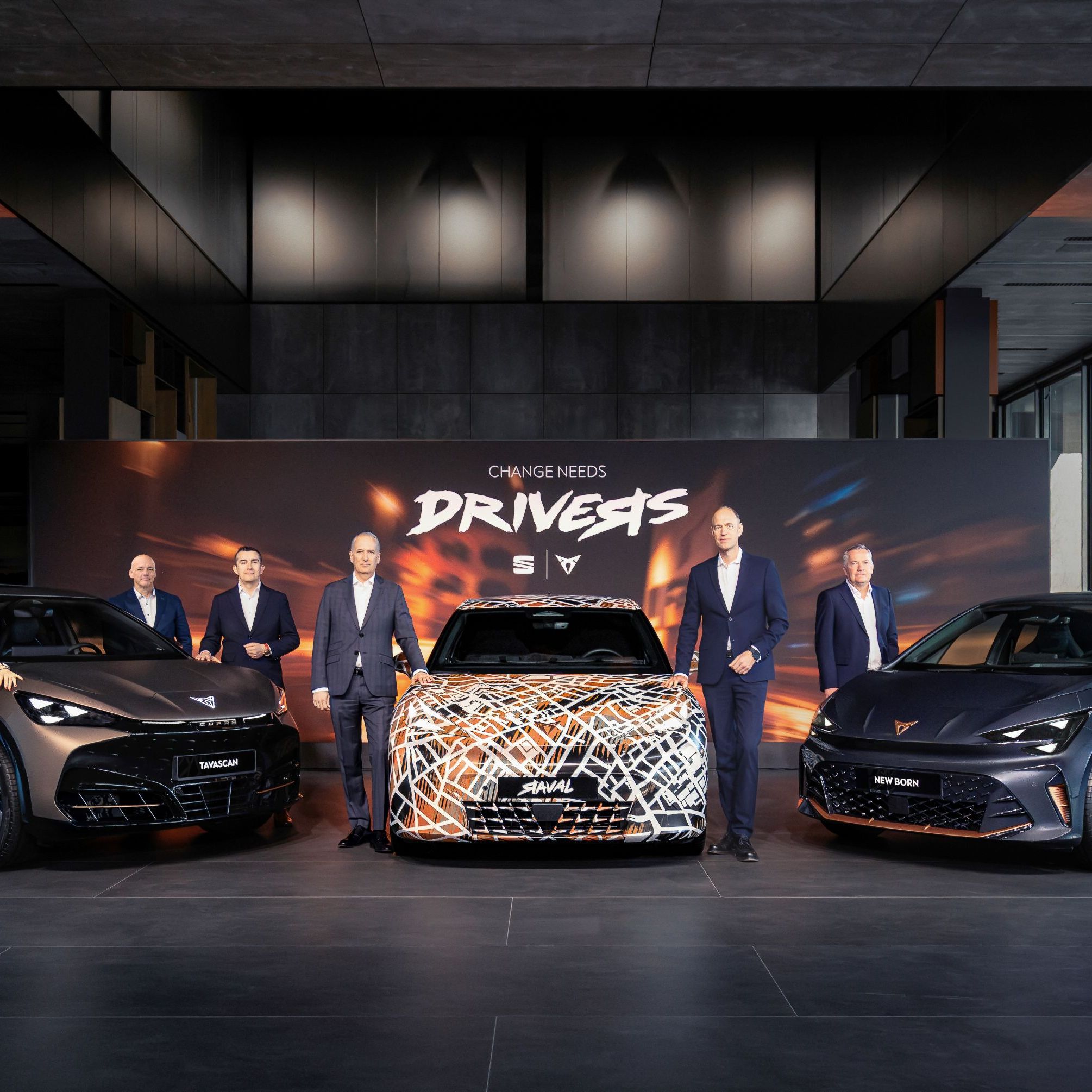 Tri modeli CUPRA zaparkované v modernom, dizajnovom showroome. Vedľa vozidiel stoja manažéri. Za vozidlami je veľký reklamný panel s nápisom "Change needs drivers." a logami SEAT a CUPRA. 