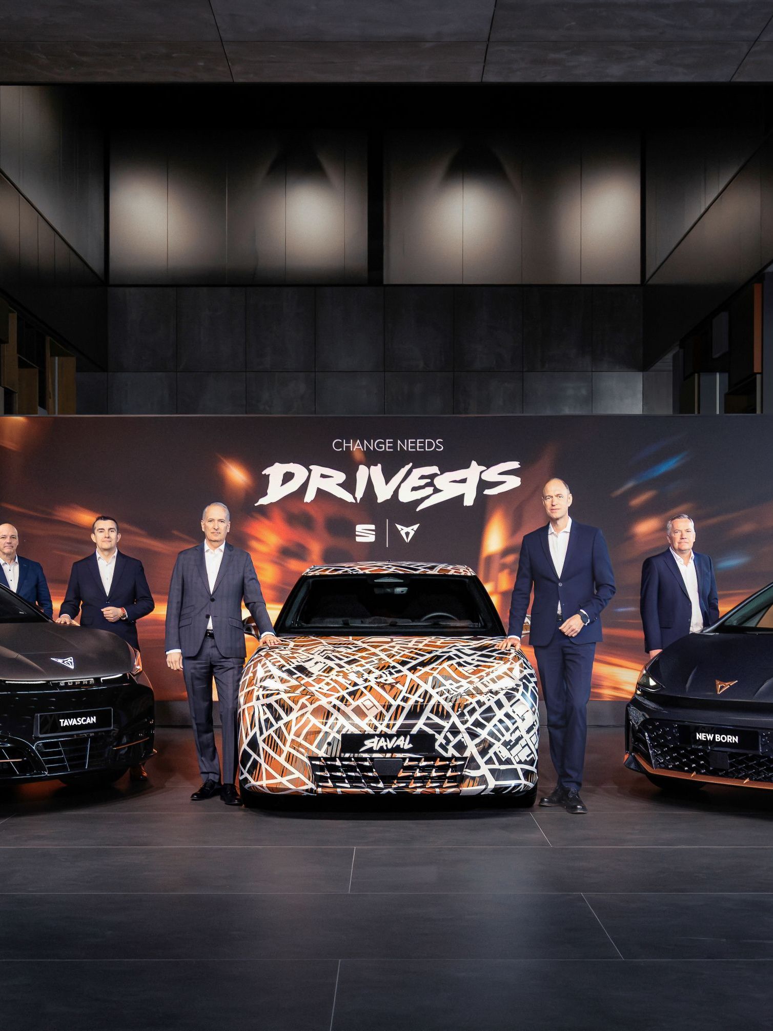 Tri modeli CUPRA zaparkované v modernom, dizajnovom showroome. Vedľa vozidiel stoja manažéri. Za vozidlami je veľký reklamný panel s nápisom "Change needs drivers." a logami SEAT a CUPRA. 