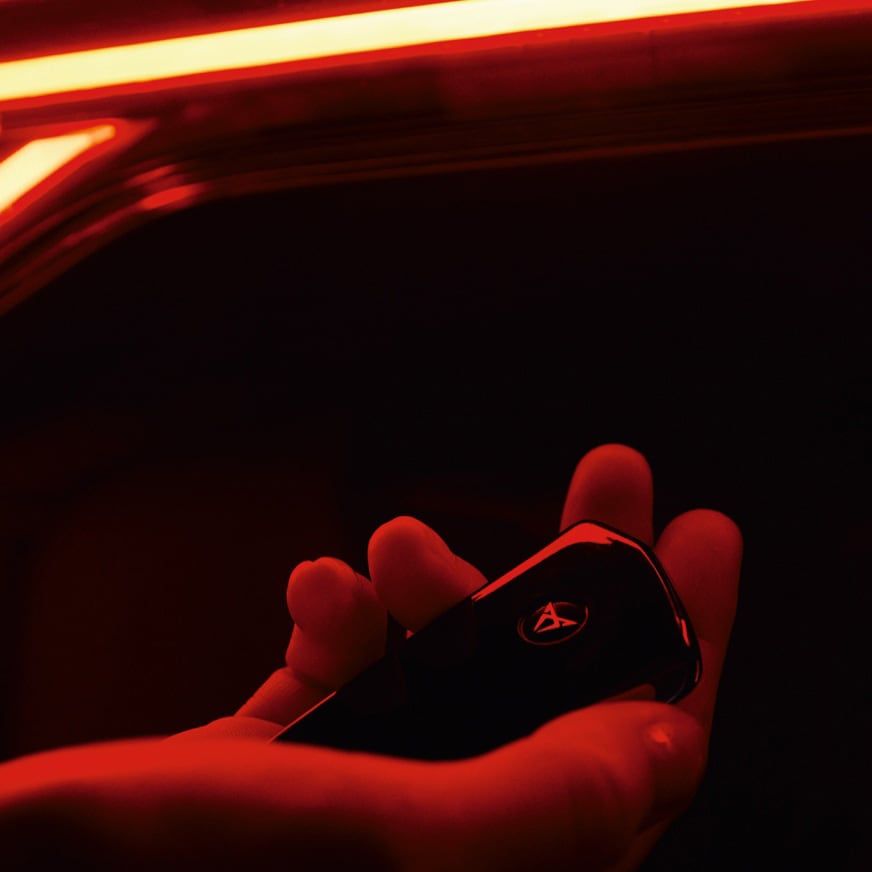 Ruka drží čierny kľúč od vozidla. Okolo je červené osvetlenie, pôsobiace dramatickým dojmom. Kľúč má logo značky.