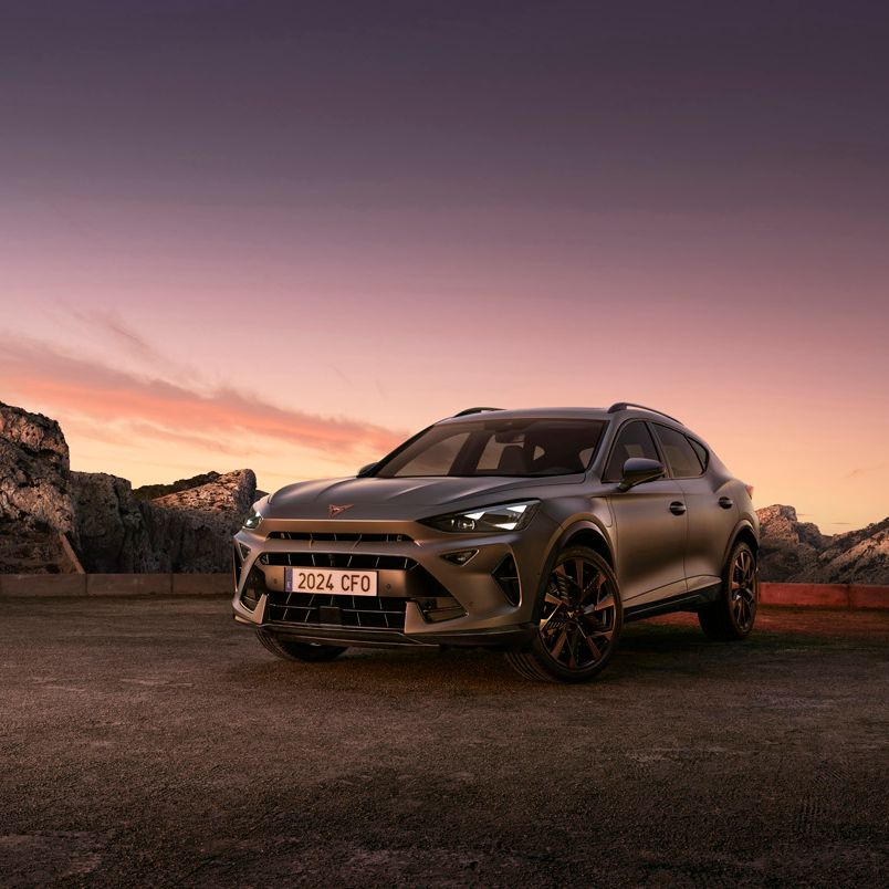 SUV športového charakteru stojí na parkovisku s výhľadom na skaly, more a západ slnka. Auto má športový dizajn a je šedej farby. Na oblohe dominuje oranžová a fialová farba pri zapadajúcom slnku.