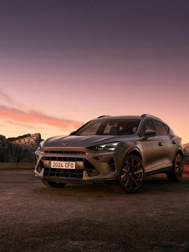 SUV športového charakteru stojí na parkovisku s výhľadom na skaly, more a západ slnka. Auto má športový dizajn a je šedej farby. Na oblohe dominuje oranžová a fialová farba pri zapadajúcom slnku.