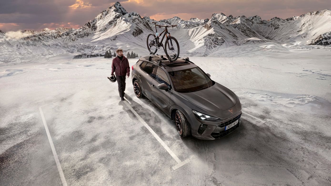 Osoba so helmou v ruke kráča vedľa šedého auta, na streche ktorého je bicykel. Scéna sa odohráva na zamrznutom a zasneženom horskom parkovisku za súmraku. V pozadí sú zasnežené hory.