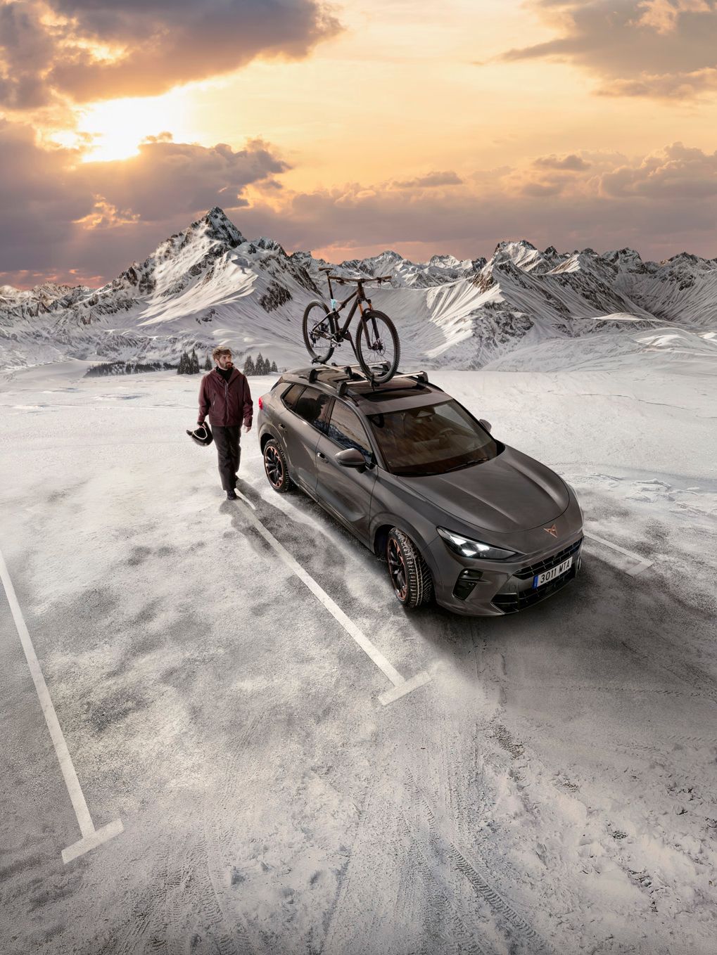 Osoba so helmou v ruke kráča vedľa šedého auta, na streche ktorého je bicykel. Scéna sa odohráva na zamrznutom a zasneženom horskom parkovisku za súmraku. V pozadí sú zasnežené hory.