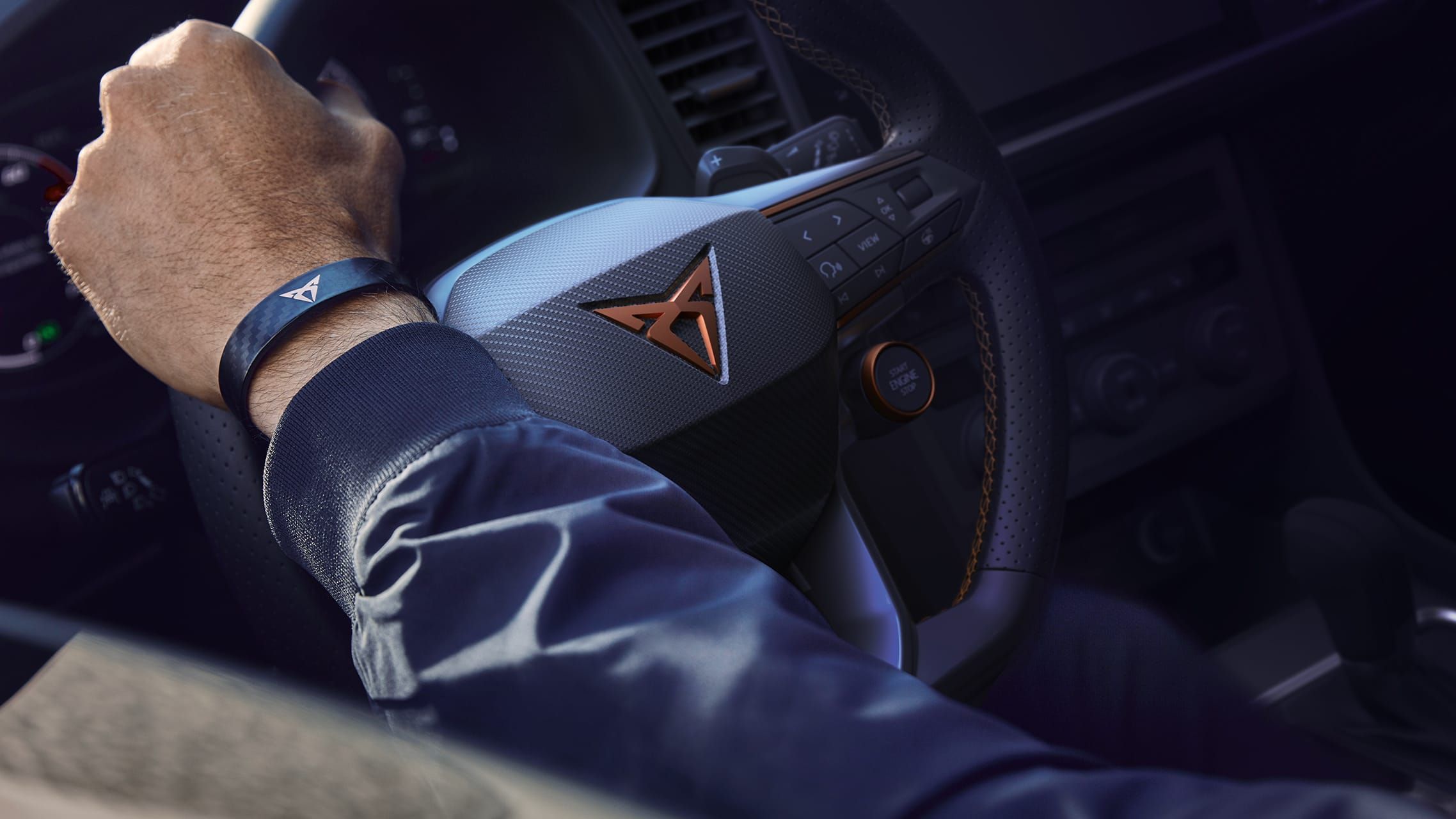 Ruka osoby, ktorá drží volant. Na ruke má náramok s logom Cupra. Interiér auta je moderný.