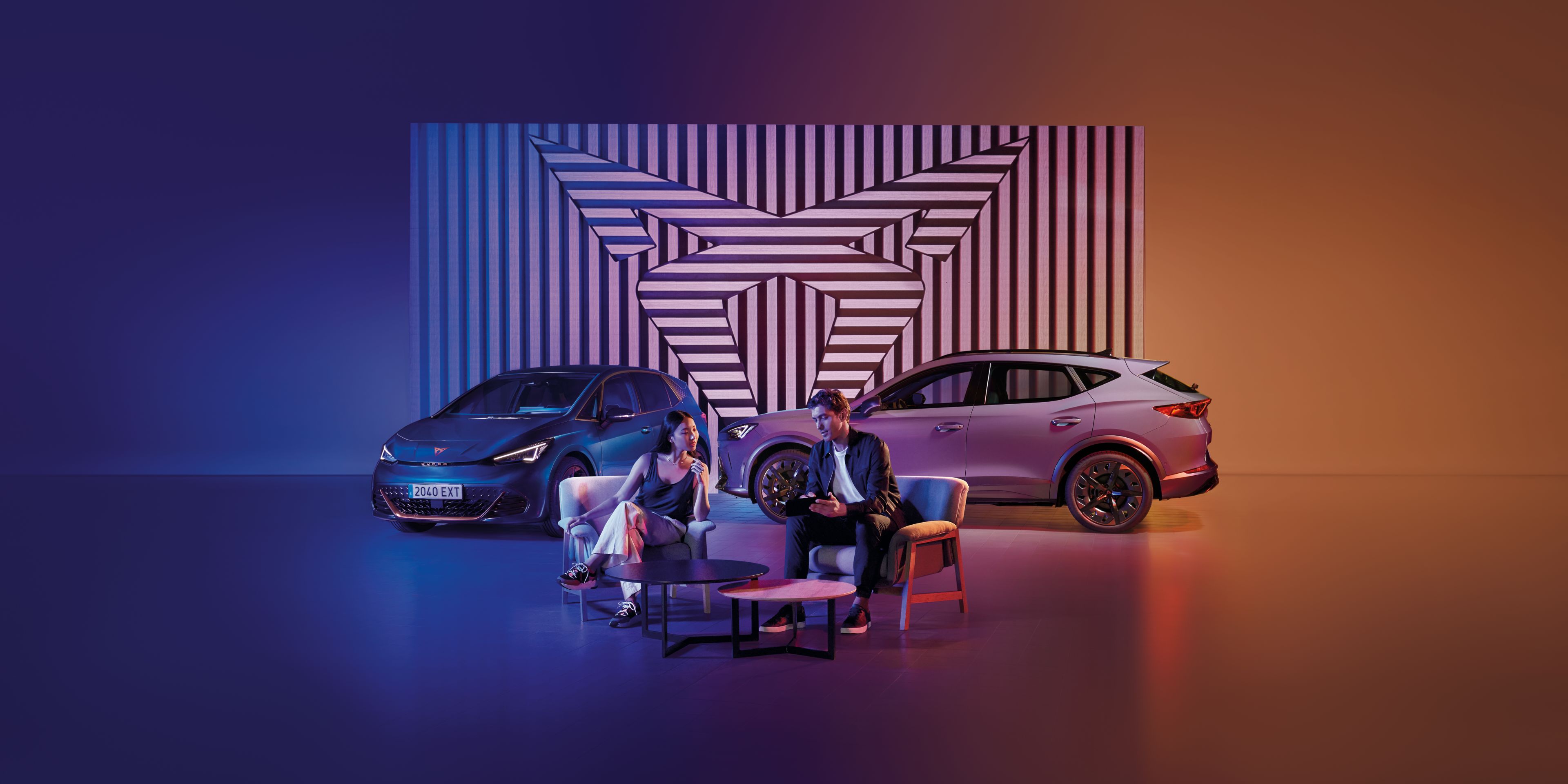 Muž a žena sedia pri okrúhlom stole na pódiu. Za nimi sú zaparkované dve autá, pozadie tvorí geometrický obraz v logom CUPRA. Scéna je osvetlená modrým a oranžovým svetlom.