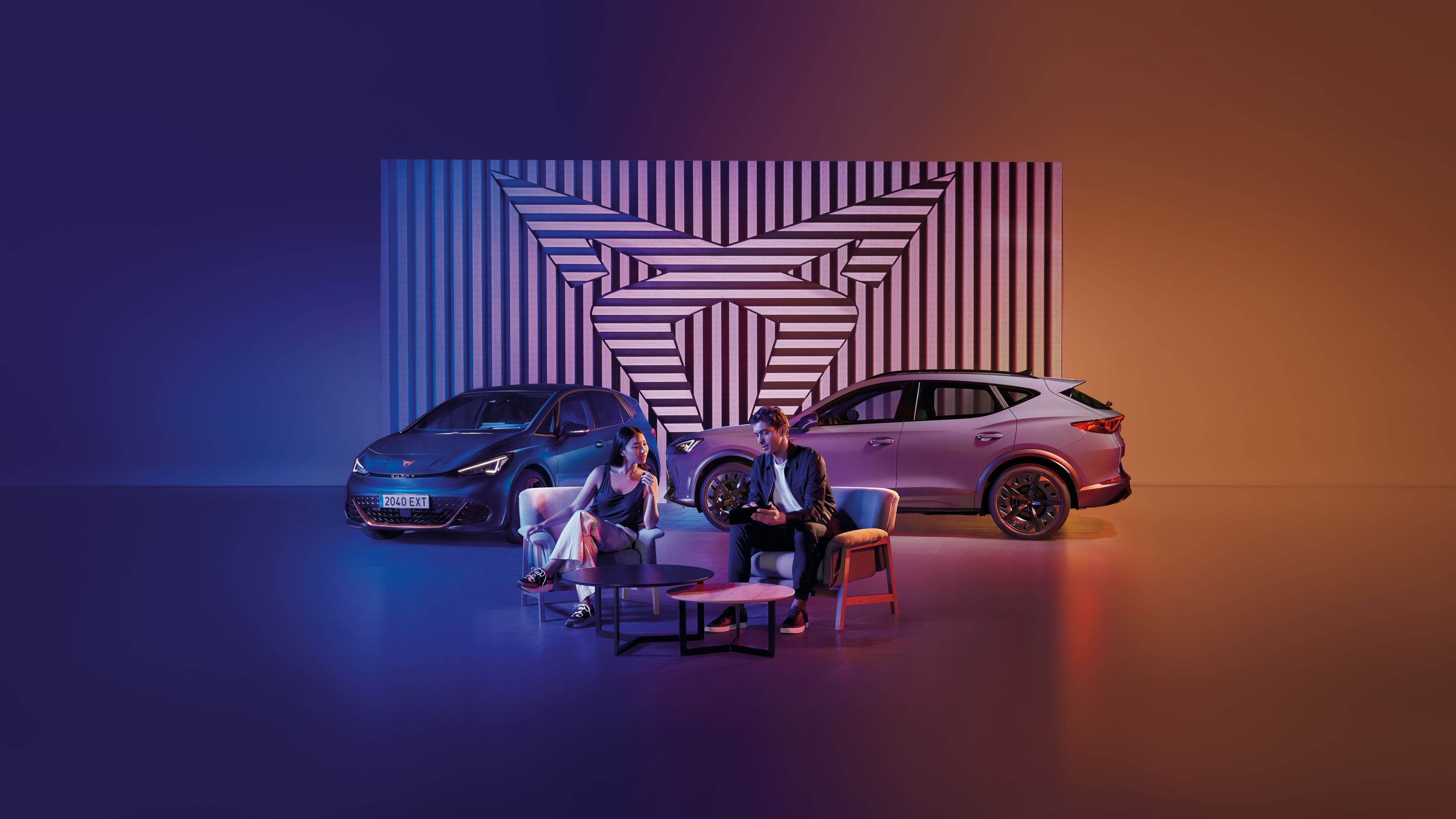 Muž a žena sedia pri okrúhlom stole na pódiu. Za nimi sú zaparkované dve autá, pozadie tvorí geometrický obraz v logom CUPRA. Scéna je osvetlená modrým a oranžovým svetlom.