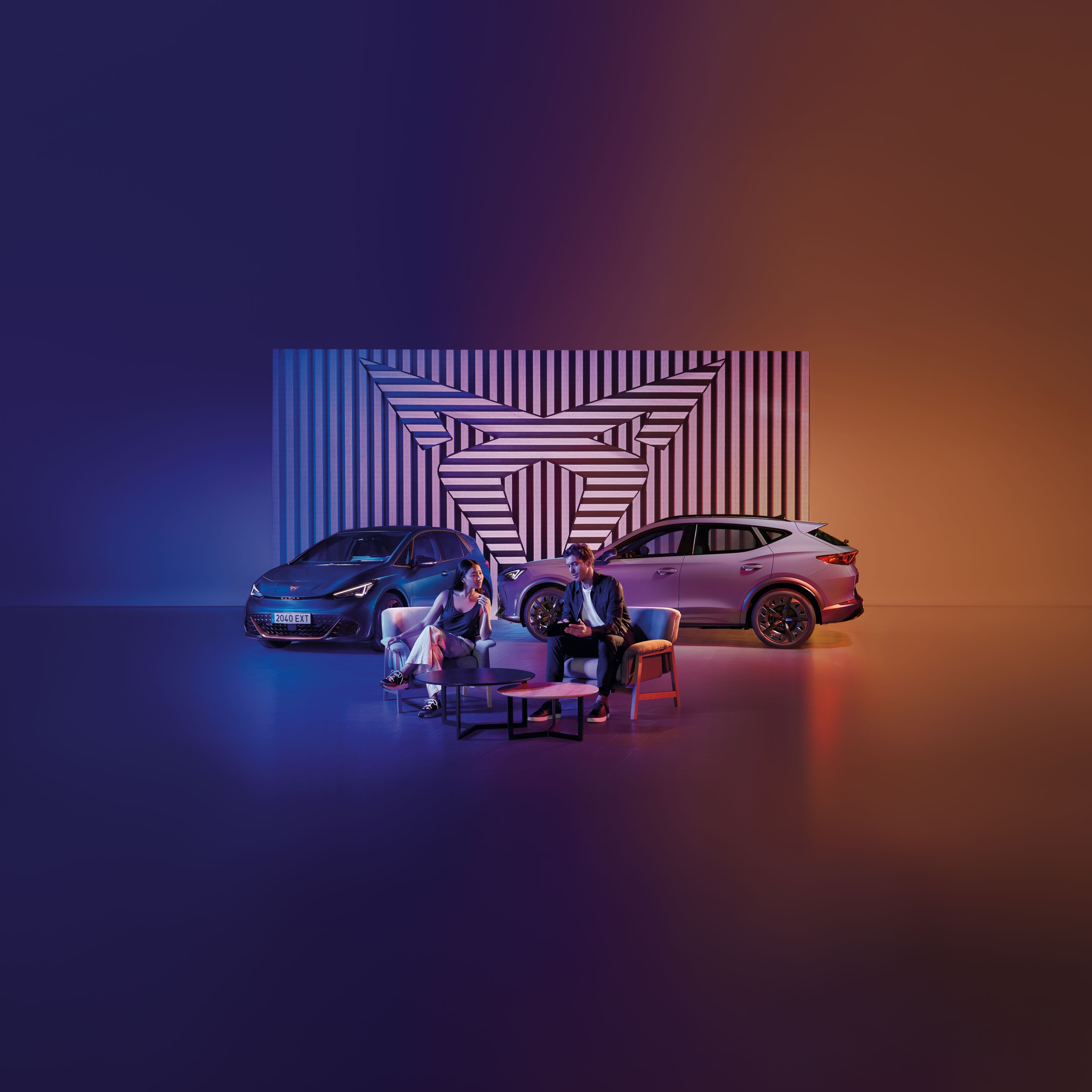 Muž a žena sedia pri okrúhlom stole na pódiu. Za nimi sú zaparkované dve autá, pozadie tvorí geometrický obraz v logom CUPRA. Scéna je osvetlená modrým a oranžovým svetlom.