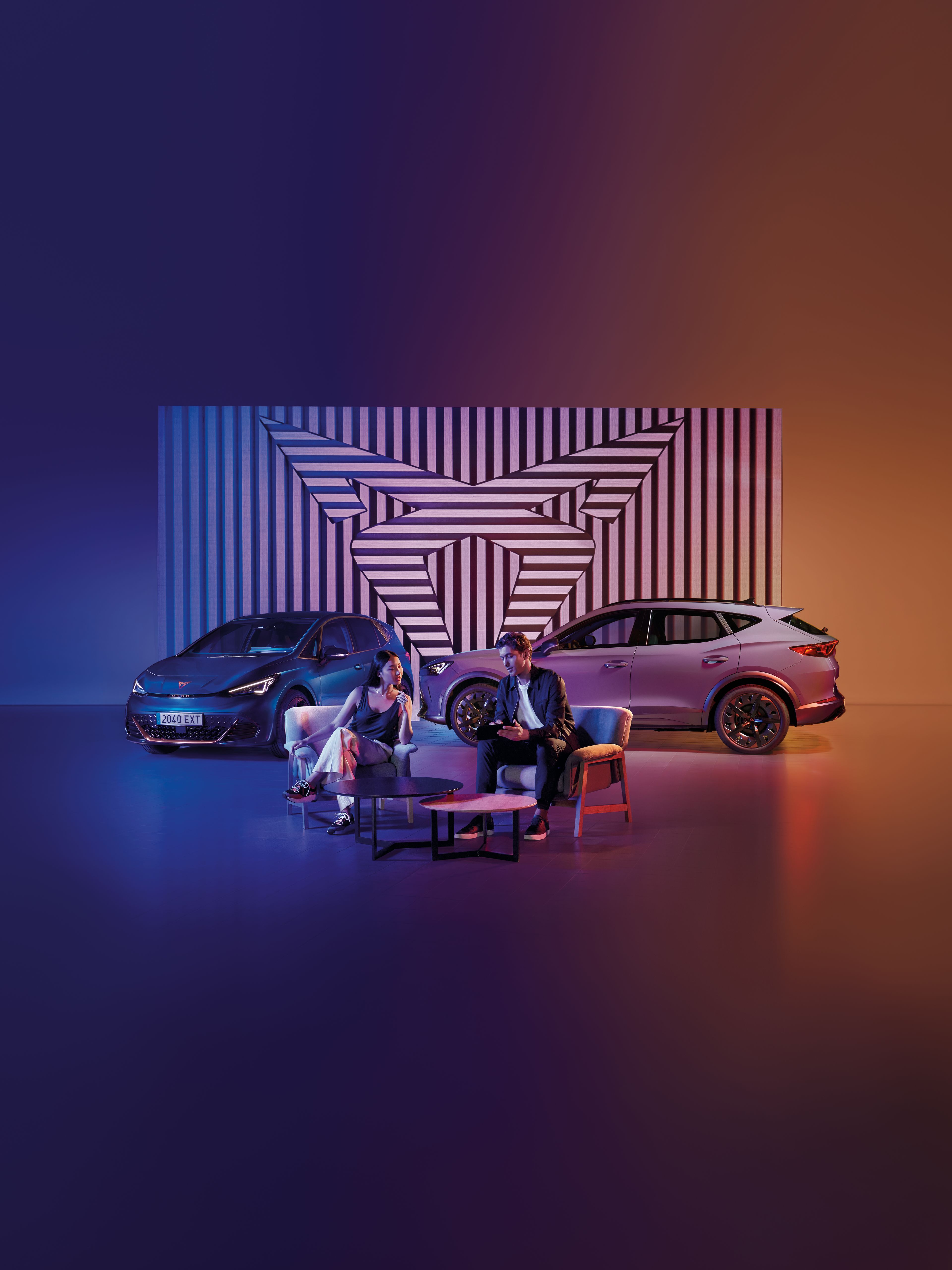 Muž a žena sedia pri okrúhlom stole na pódiu. Za nimi sú zaparkované dve autá, pozadie tvorí geometrický obraz v logom CUPRA. Scéna je osvetlená modrým a oranžovým svetlom.