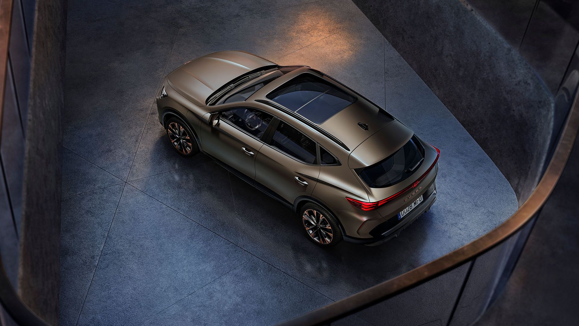 Moderné hnedé SUV je zaparkované v tmavej miestnosti, pohľad zhora ukazuje jeho elegantné línie. Hladký povrch vozidla zdôrazňuje jeho moderný dizajn. Široké okná dávajú interiéru svetlý a vzdušný pocit.