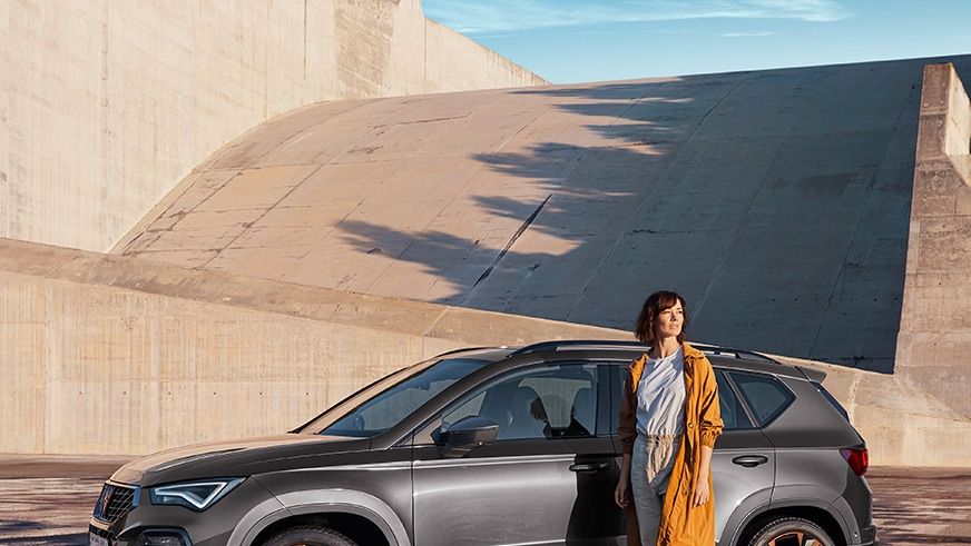 Osoba v oranžovom kabáte stojí vedľa sivého SUV, ktoré je zaparkované na mokrom povrchu. V pozadí je betónová stena a modrá obloha.