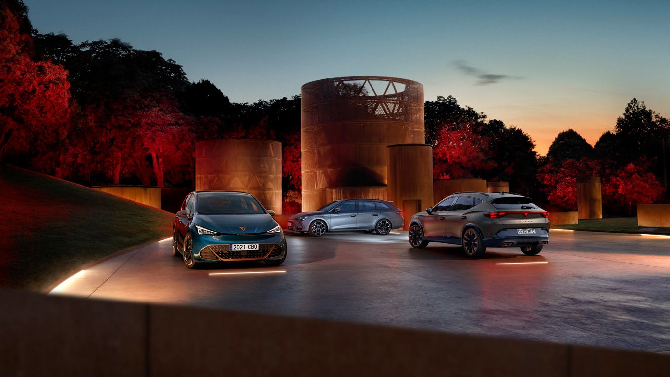 Tri modely CUPRA stoja večer pri zapadajúcom slnku na osvetlenej ploche pri moderných stavbách v tvare valca, za ktorými je hustý park.