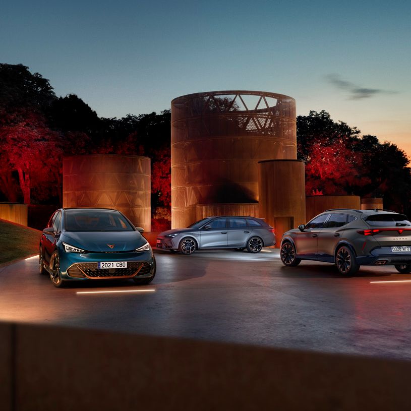 Tri modely CUPRA stoja večer pri zapadajúcom slnku na osvetlenej ploche pri moderných stavbách v tvare valca, za ktorými je hustý park.