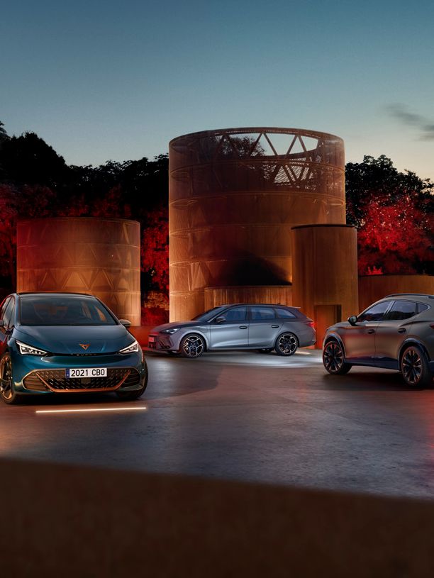 Tri modely CUPRA stoja večer pri zapadajúcom slnku na osvetlenej ploche pri moderných stavbách v tvare valca, za ktorými je hustý park.